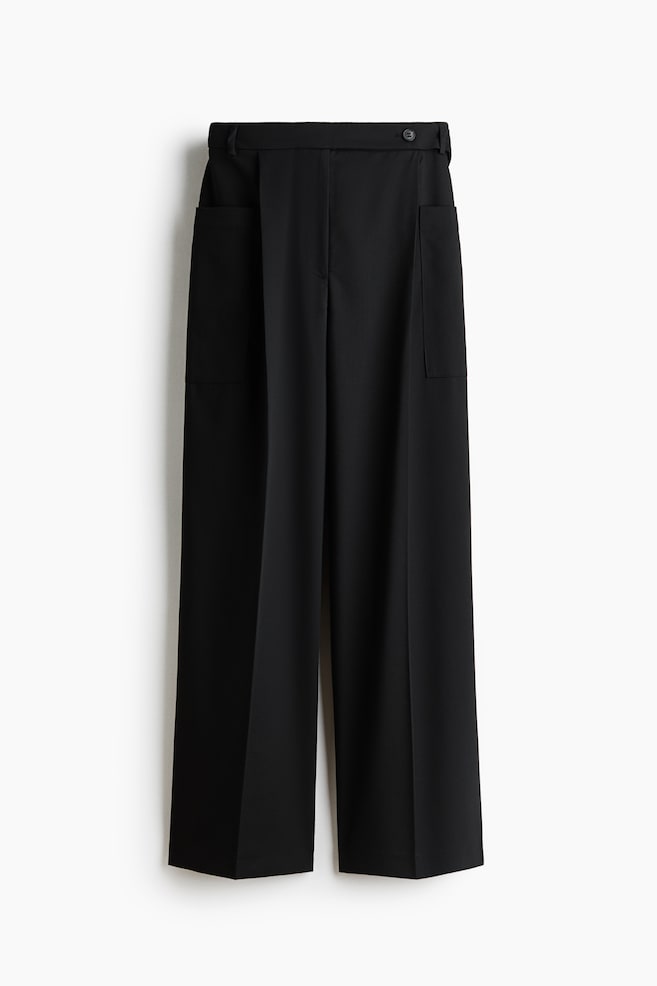 A pair of black wide-leg trousers is hanging front-facing, featuring a structured waistband with belt loops and an asymmetrical button fastening. The trousers display sharp pressed creases down the centre of each leg and deep patch pockets at the sides. - 1