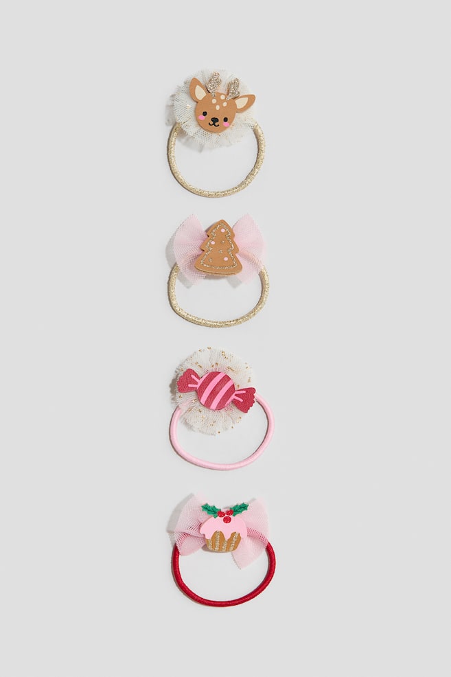 Four festive hair ties are presented in a vertical line from a front view, each featuring a unique decorative holiday element. The designs include a reindeer head with glittery antlers on a gold band, a gingerbread tree on a pink tulle bow with a gold band, a pink wrapped candy on a speckled white tulle pom-pom with a pink band, and a holiday cupcake with holly on a pink tulle bow with a red band. - 1
