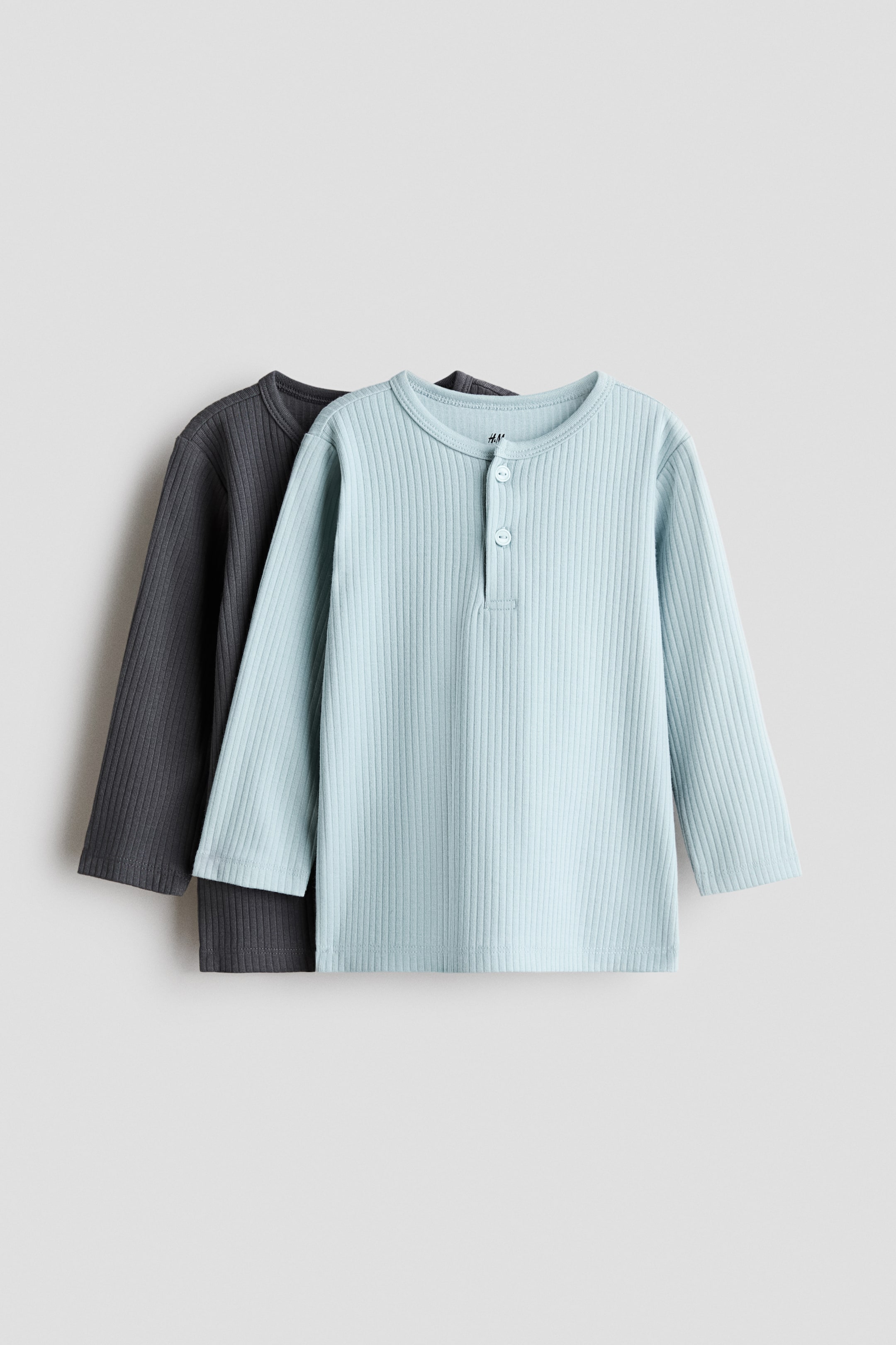 View larger image: Two long-sleeved tops are shown facing front, both featuring a soft, ribbed cotton jersey texture. The light blue top is positioned in front, displaying a round, trimmed neckline and a three-button placket, with a dark grey top layered behind it.