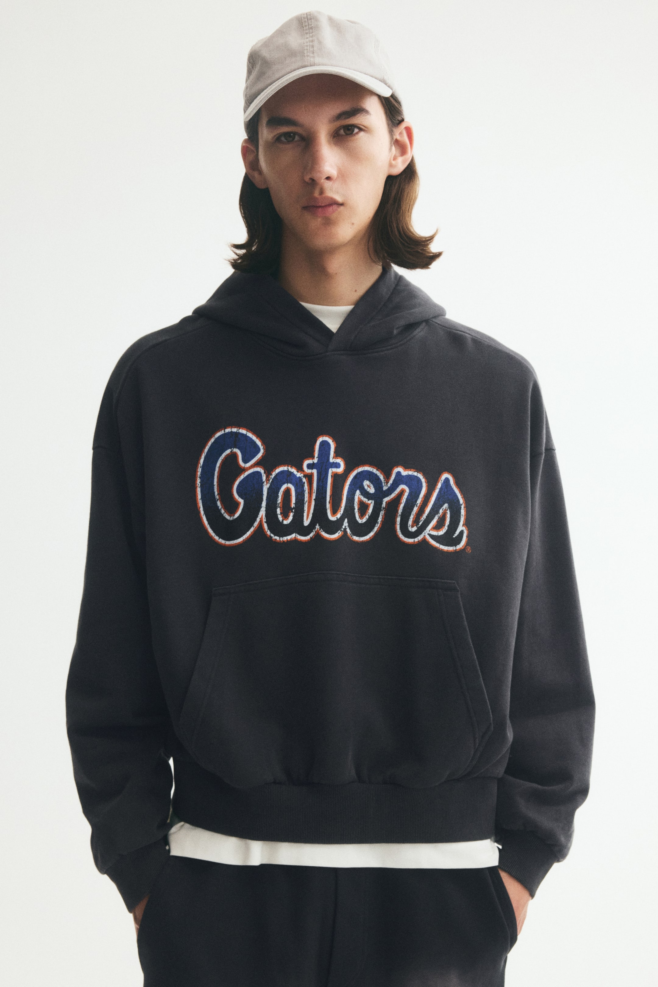 Grotere afbeelding bekijken: Een donkergrijze oversized hoodie met een verweerde blauwe en oranje "Gators"-scriptprint op de voorkant wordt gedragen door het model. De hoodie is ontworpen met een omlijnde capuchon, verlaagde schouders, een kangoeroezak en geribde boorden, en is gestyled met een gelaagd wit T-shirt eronder en een lichtgrijze pet.
