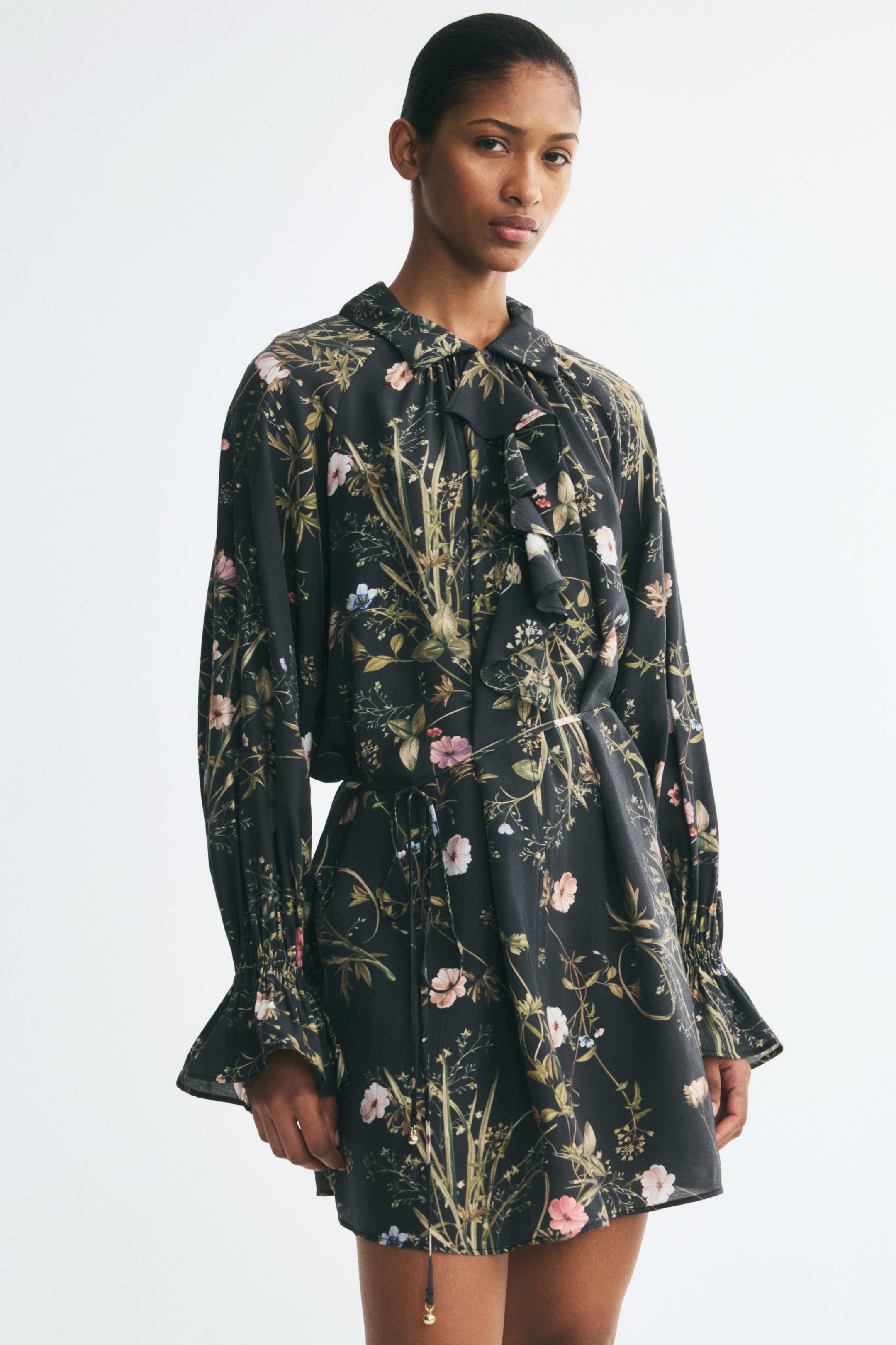 View larger image: A short floral-patterned dress in dark fabric is worn by the model, featuring a buttoned collar with a wide chest flounce and a slim tie belt at the waist. The dress has long balloon sleeves with elasticated wrists and flared cuffs, flowing into a gently flared skirt.