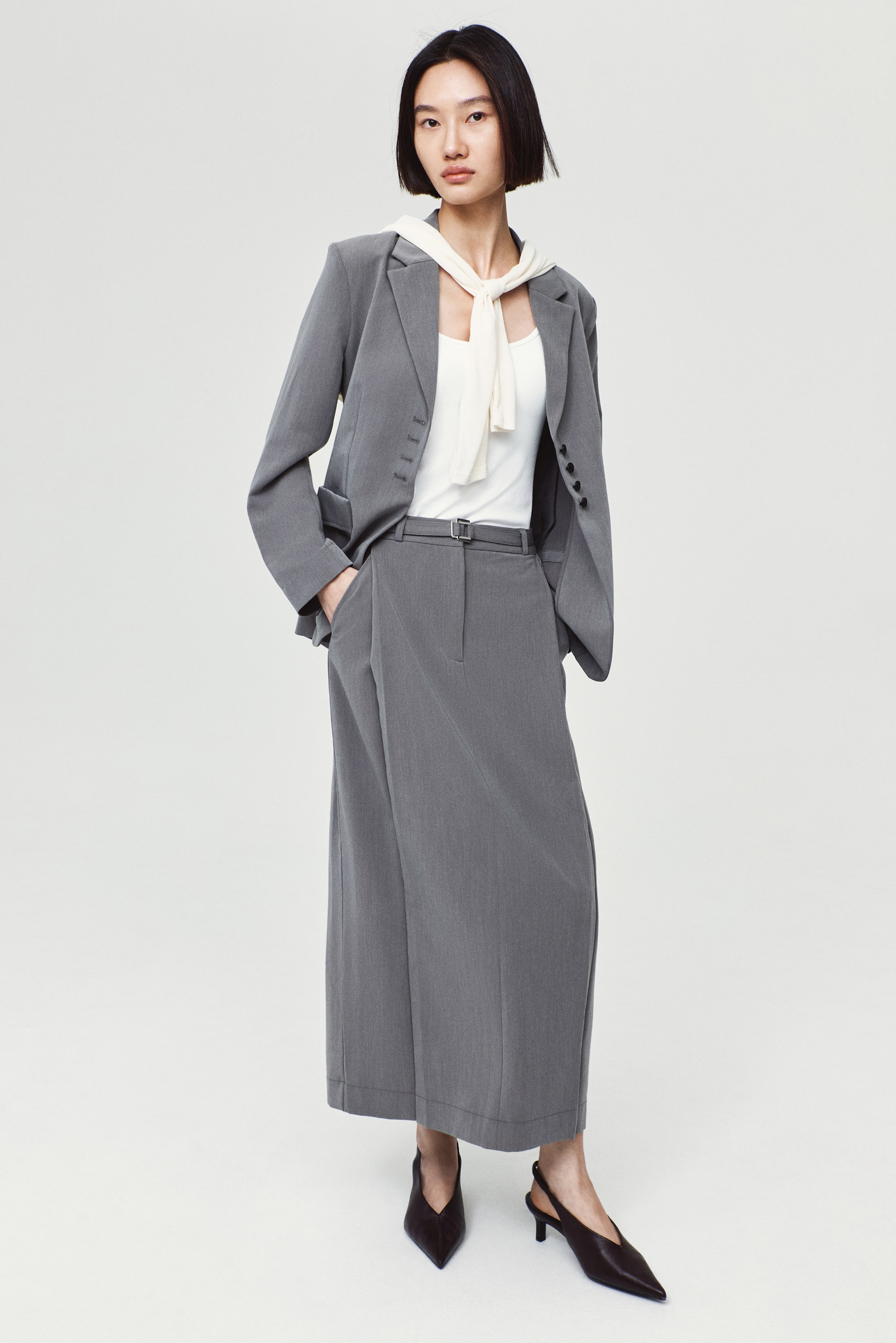 View larger image: A calf-length grey twill skirt featuring front pleats and a thin belt with a silver buckle is worn by the model, who stands with one hand in its diagonal pocket. The skirt is styled as a suit with a matching grey blazer, a white top with a tied cream scarf, and black slingback heels.