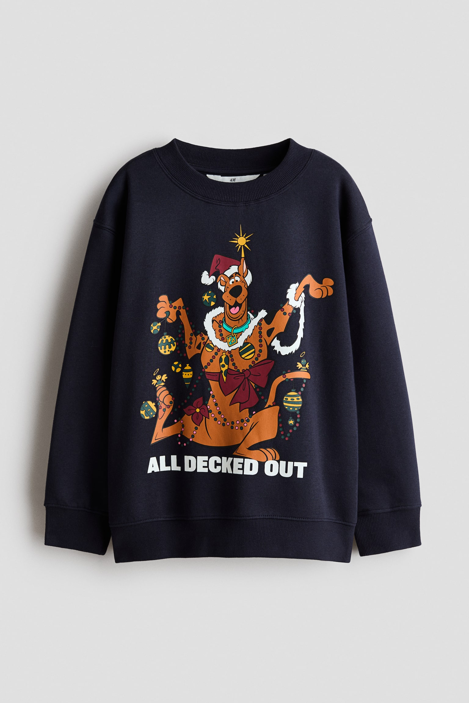 Se muestra de frente una sudadera holgada en azul marino oscuro, con cuello redondo acanalado, hombros caÃdos y puños y bajo acanalados. En el centro presenta un gráfico impreso del personaje de dibujos animados Scooby-Doo decorado con luces y adornos festivos, con el texto "ALL DECKED OUT" impreso en letras blancas en negrita debajo.