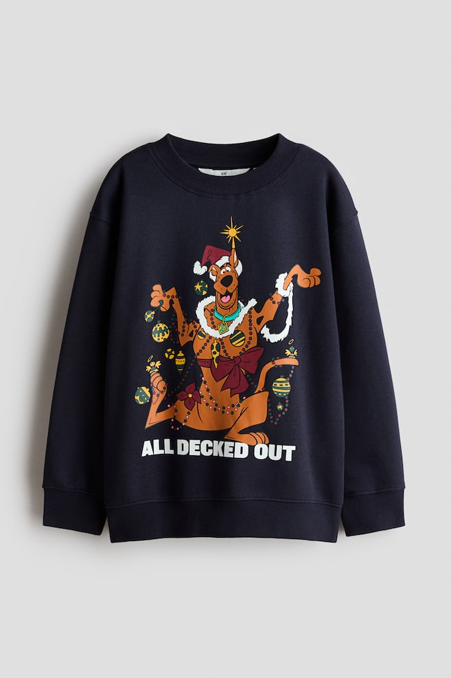 A dark navy-blue loose-fit sweatshirt is shown from the front, featuring a round rib-trimmed neckline, dropped shoulders, and ribbed cuffs and hem. The centre displays a printed graphic of the cartoon character Scooby-Doo decorated with festive lights and ornaments, with the text "ALL DECKED OUT" printed in bold white letters below. - 1