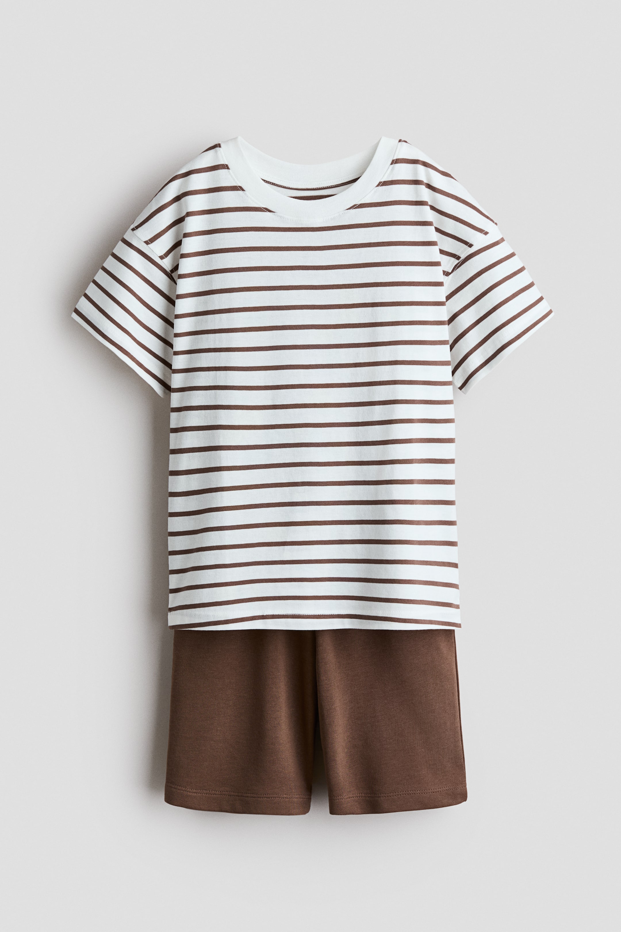Ver imagen más grande: Un conjunto se presenta desde el frente, compuesto por una camiseta de suave jersey de algodón y unos pantalones cortos de tejido ligero tipo sudadera. La camiseta es de color blanco con rayas horizontales marrones, presenta un cuello redondo acanalado, hombros caídos y un dobladillo recto. Los pantalones cortos marrones cuentan con una cintura elástica y bolsillos laterales diagonales.