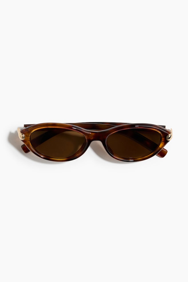 Oval sunglasses with tortoiseshell-patterned plastic frames and dark brown tinted lenses are lying flat in a front-facing view. Small gold-tone metal details accent the outer hinges near the folded temples. - 1