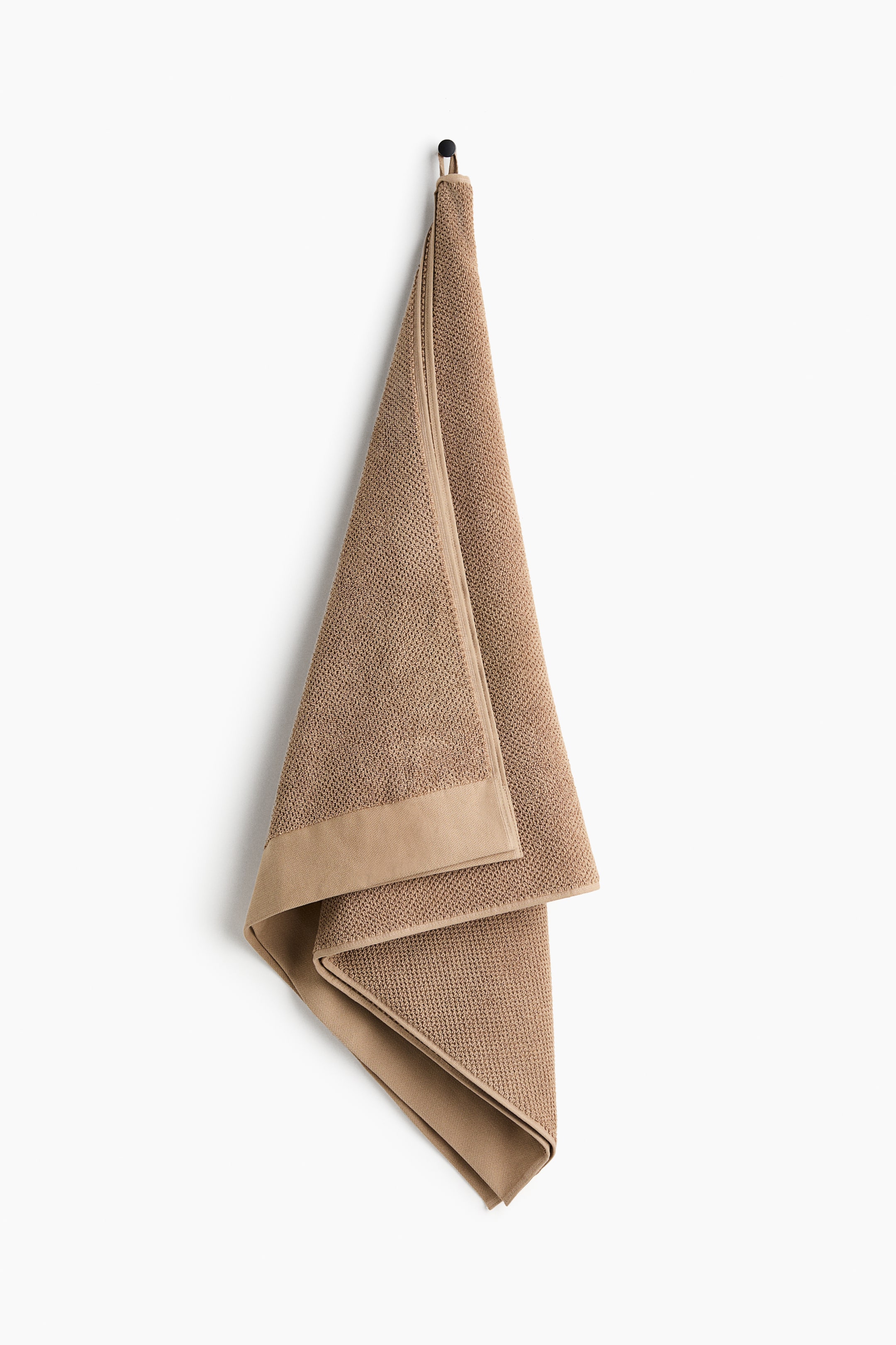 View larger image: A brown bath towel is hanging folded from a black hook, displaying a textured weave across its main body and a smooth border along one edge, with a small loop visible at the top for hanging.