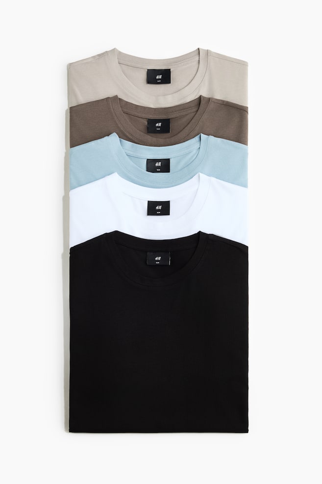 A stack of five slim-fit cotton jersey T-shirts is shown from the front, featuring round, rib-trimmed necklines. The folded T-shirts display a colour palette of light beige, brown, light blue, white, and black. - 1