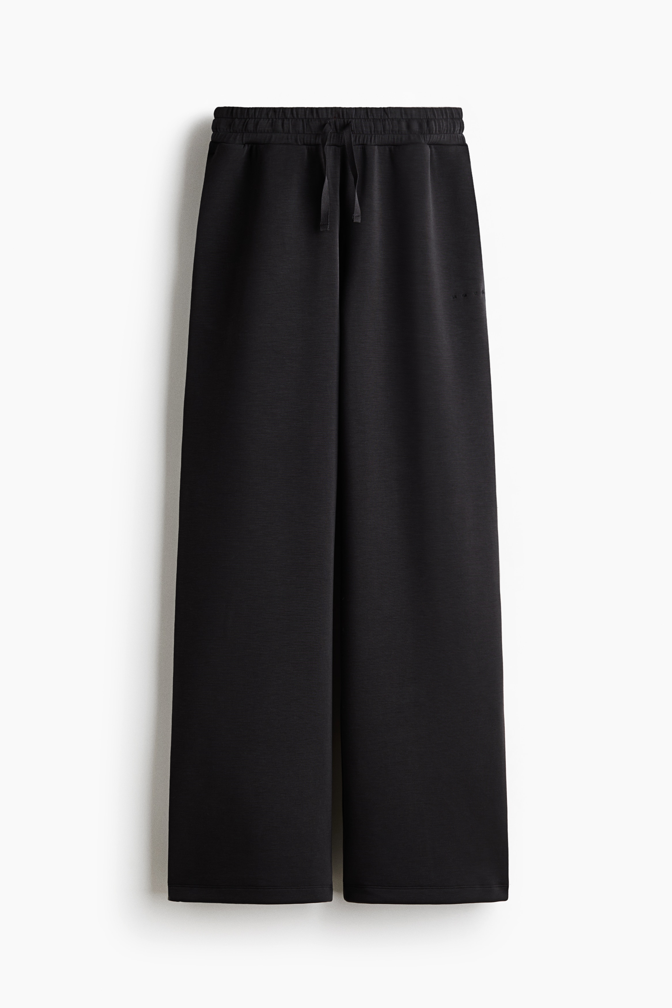 Schwarze, oversized Activewear-Jogginghosen mit weitem Bein und elastischem Bund werden vom Model getragen. Die Jogginghosen sind mit einem figurbetonten schwarzen Rollkragen-Longsleeve kombiniert, und das Model steht mit einer Hand in der Seitentasche.