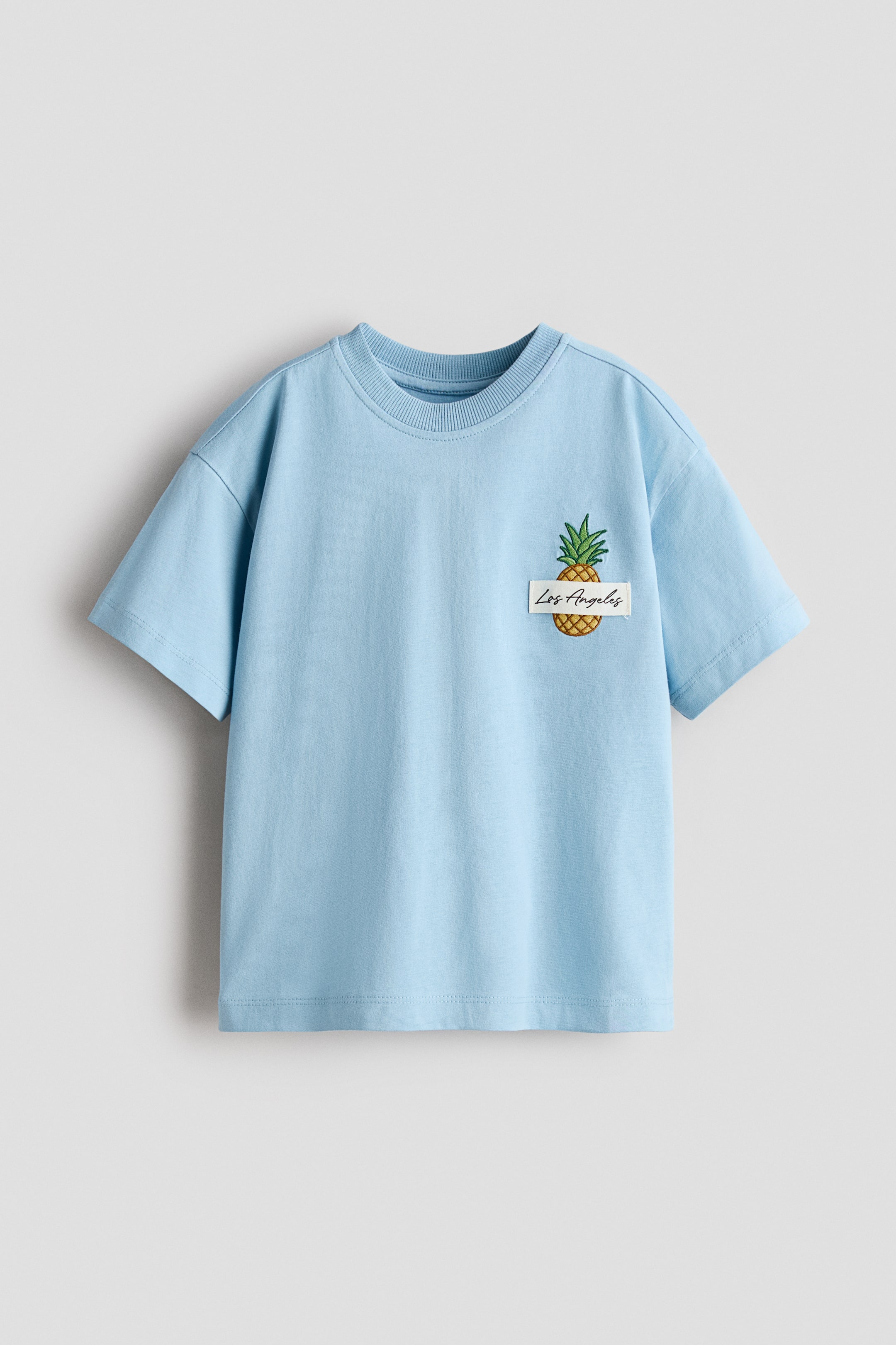 Agrandir l'image: Un t-shirt bleu clair à coupe ample est présenté de face, avec un col rond à bordure côtelée, des épaules tombantes et des manches courtes. Un motif imprimé sur le devant affiche un ananas avec une étiquette rectangulaire blanche superposée, présentant "Los Angeles" en texte manuscrit.