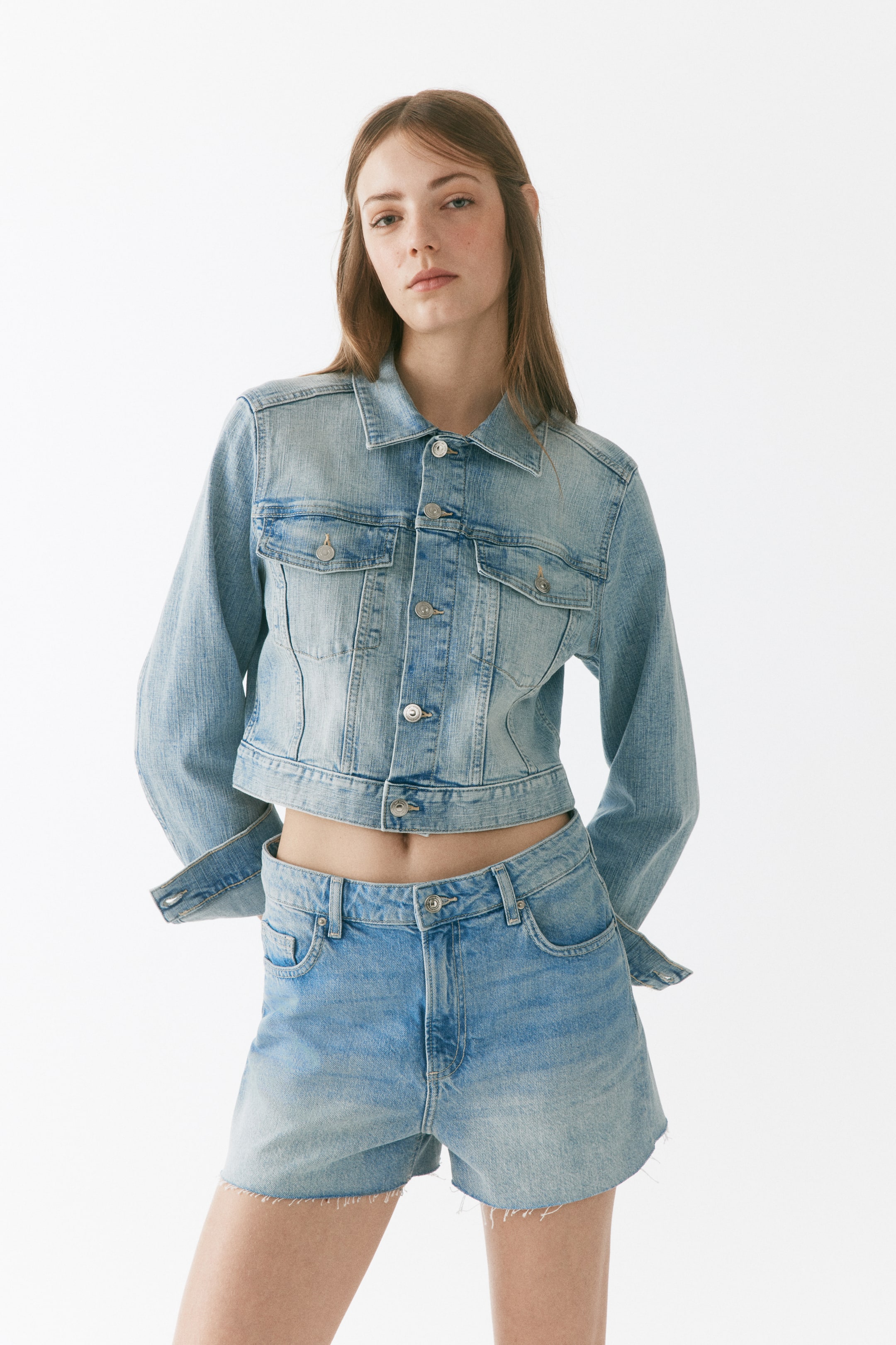 Größeres Bild ansehen: Eine hell gewaschene, gekürzte Jeansjacke wird getragen, nach vorne gerichtet, mit einem klassischen Kragen, einem vollständigen Knopfverschluss vorne, zwei Brusttaschen mit Klappen und langen Ärmeln mit Knopfmanschetten.