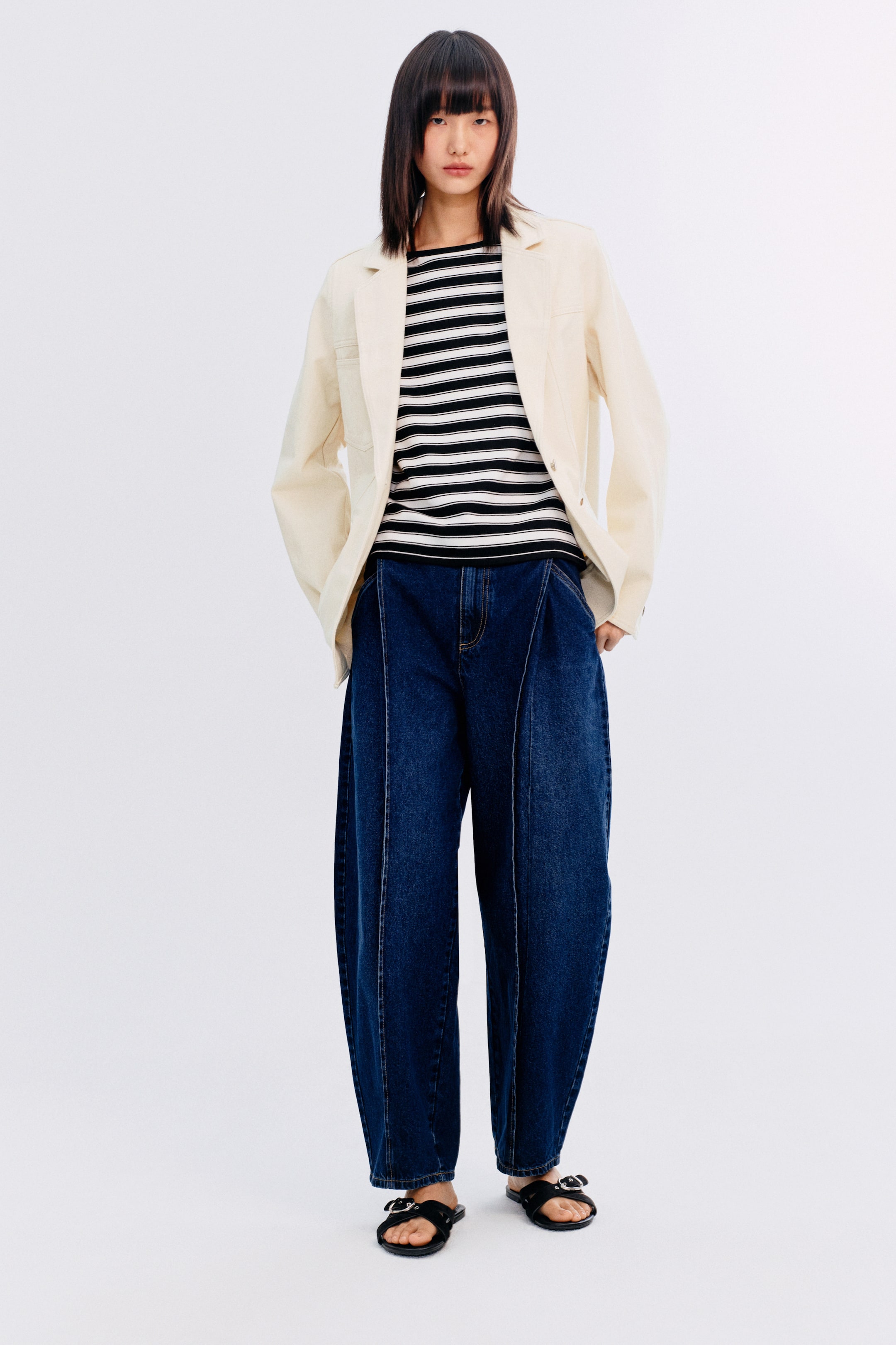 拡大画像を表示: オフホワイトのジャケットが、黒と白のストライプの長袖トップの上にオープンに着用されています。濃い青のワイドレッグジーンズは、目立つ縦のフロントシームがあり、黒のバックルサンダルと組み合わせられています。