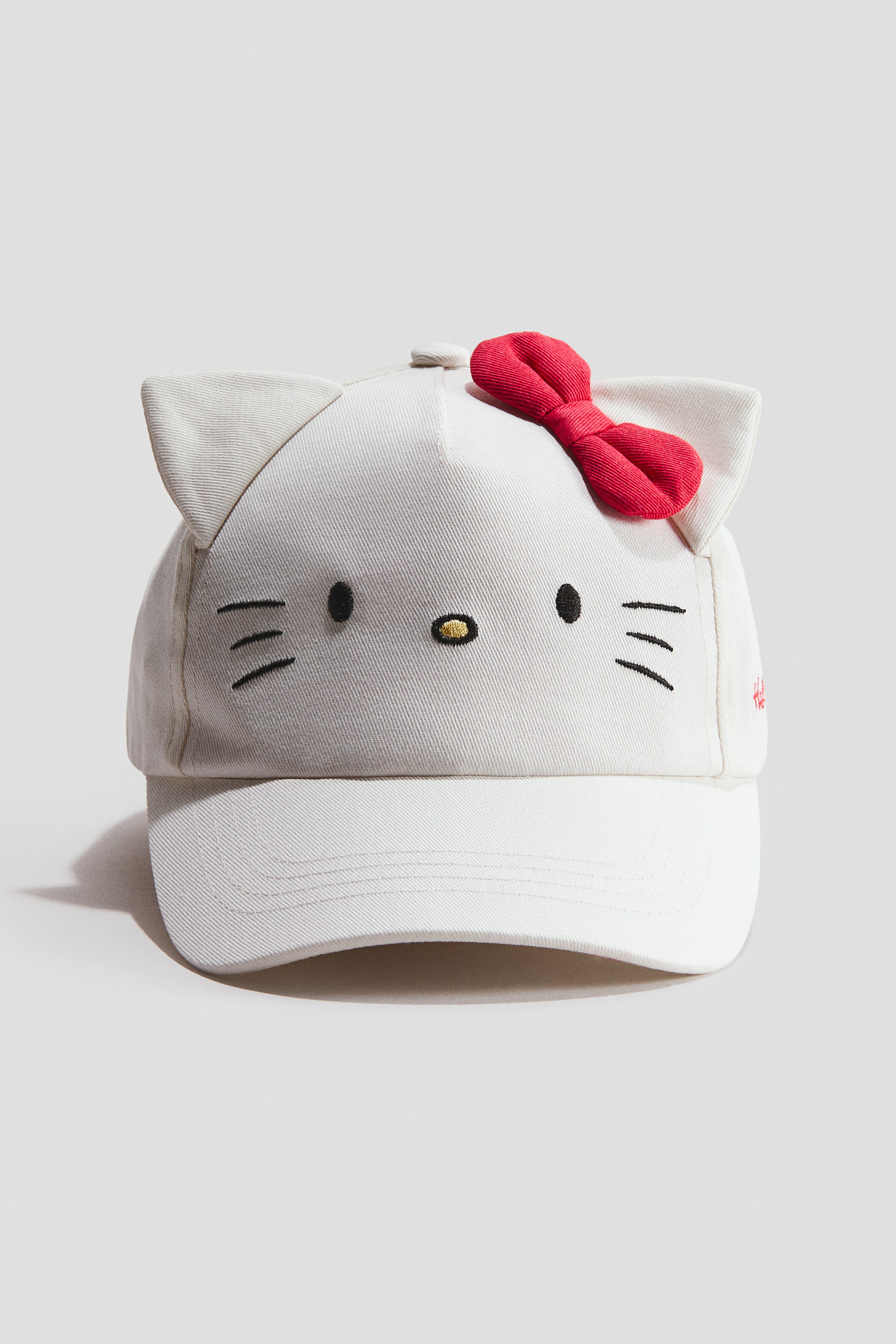 View larger image: A white baseball cap with prominent cat ears perched on top and a red bow adorning the right side. The front showcases an embroidered cat face with two black oval eyes, a yellow oval nose, and black whiskers, while a small red embroidered text is visible on the cap's lower right side.