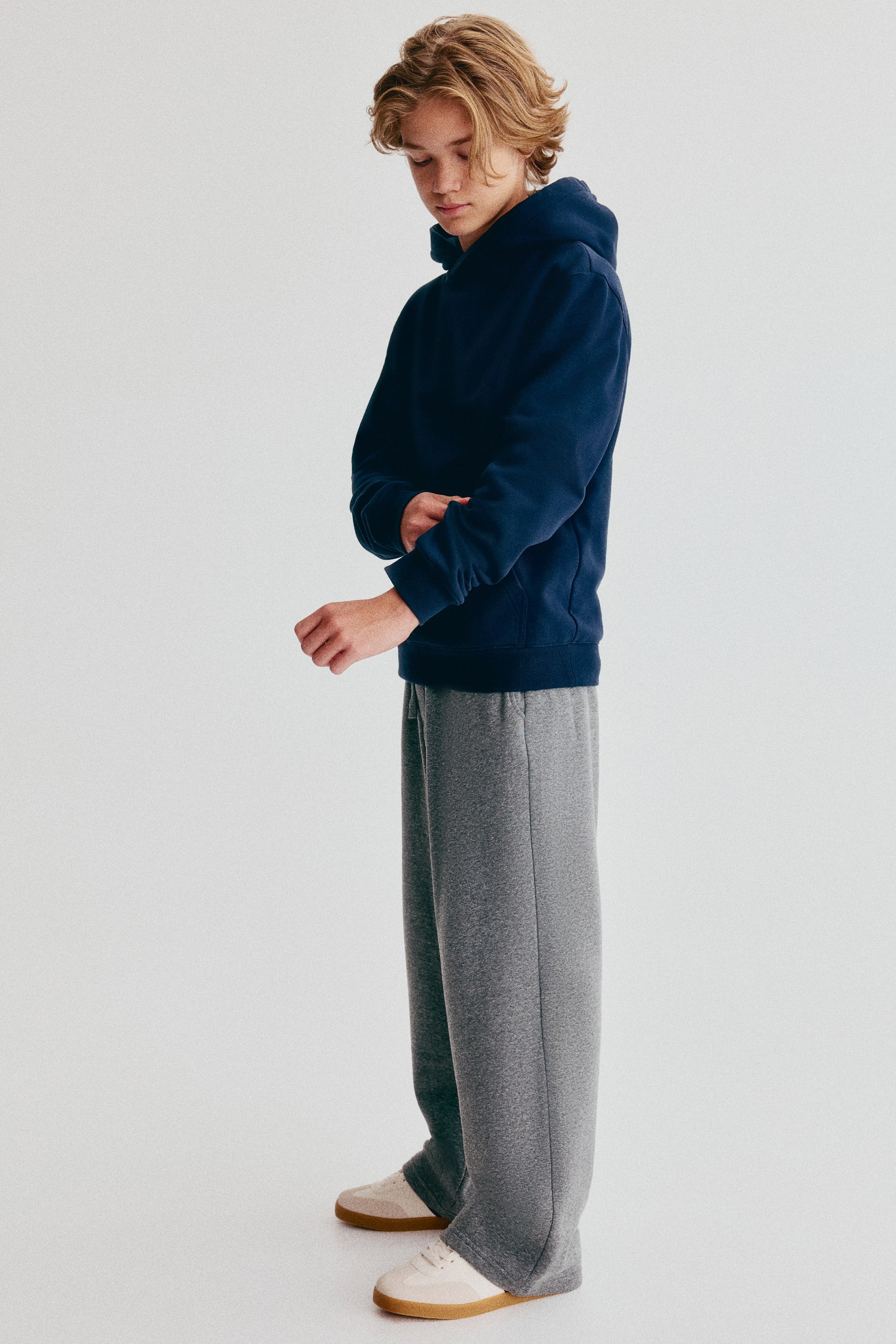 Vezi imaginea mai mare: Pantalonii de trening gri sunt prezentați din profil lateral, orientați spre stânga, având o croială wide-leg și o potrivire lejeră. Pantalonii cad relaxat peste pantofi sport albi și sunt asortați cu un hanorac bleumarin pentru a evidenția aspectul lor casual.