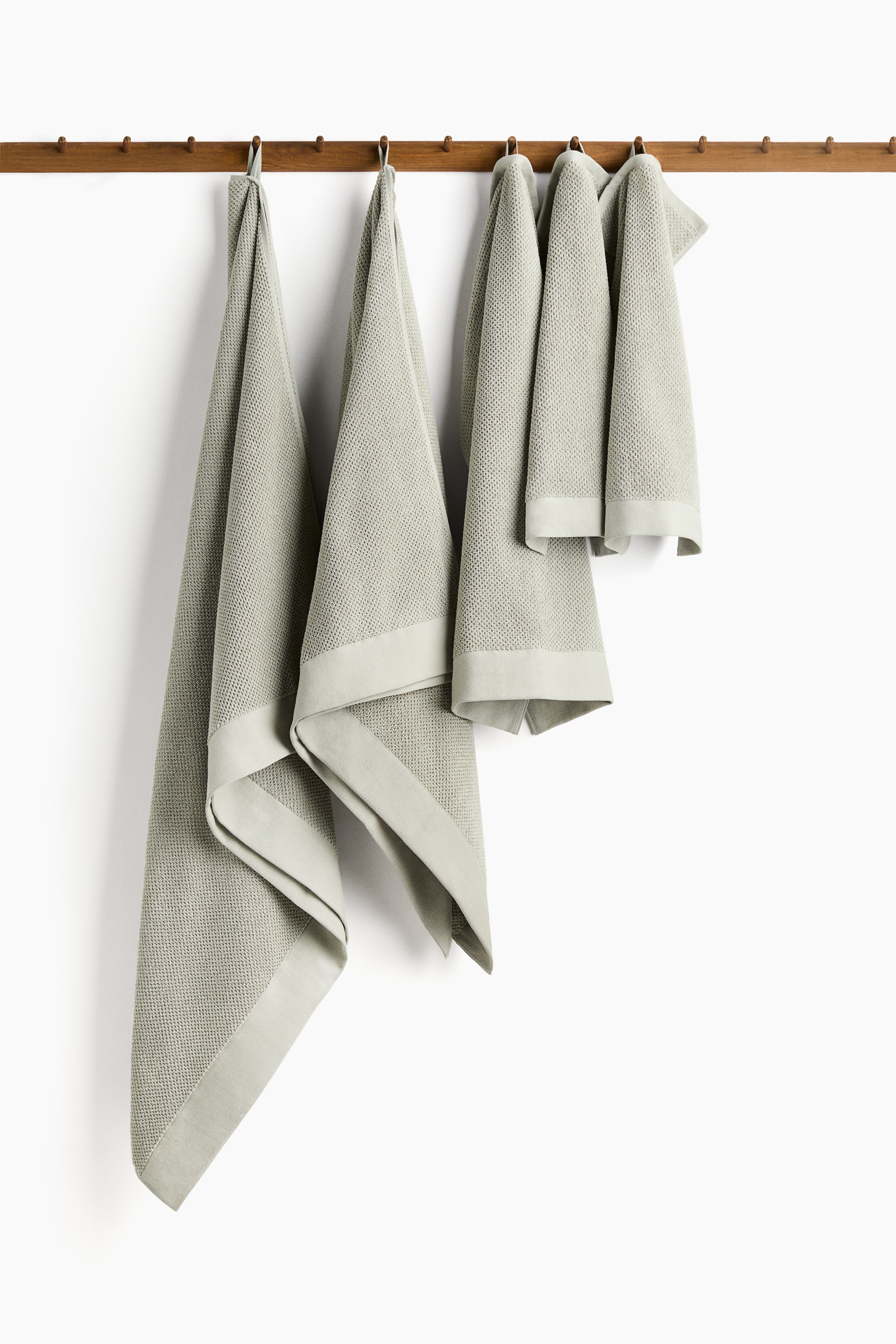 View larger image: Four light green towels of different sizes are hanging from hooks on a wooden rail, displaying a subtle textured weave and smooth, flat borders. Each towel is featuring a hanging loop at its top edge.