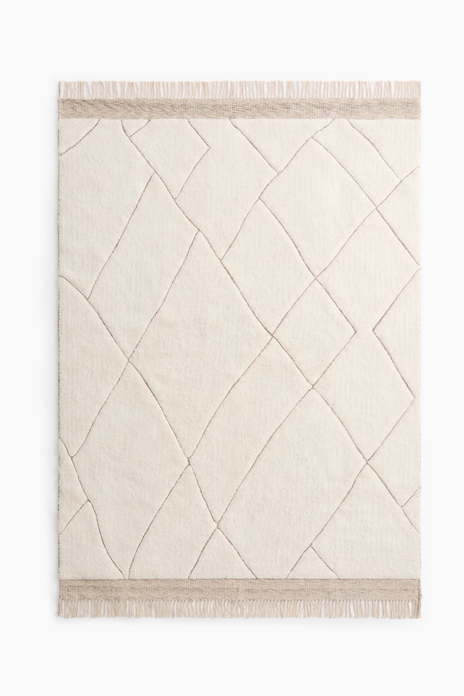 Un tapis rectangulaire blanc cassé présente un motif géométrique irrégulier avec des lignes surélevées de couleur brun clair, et des bandes tissées avec des franges apparaissent le long de ses bords supérieur et inférieur. - 1