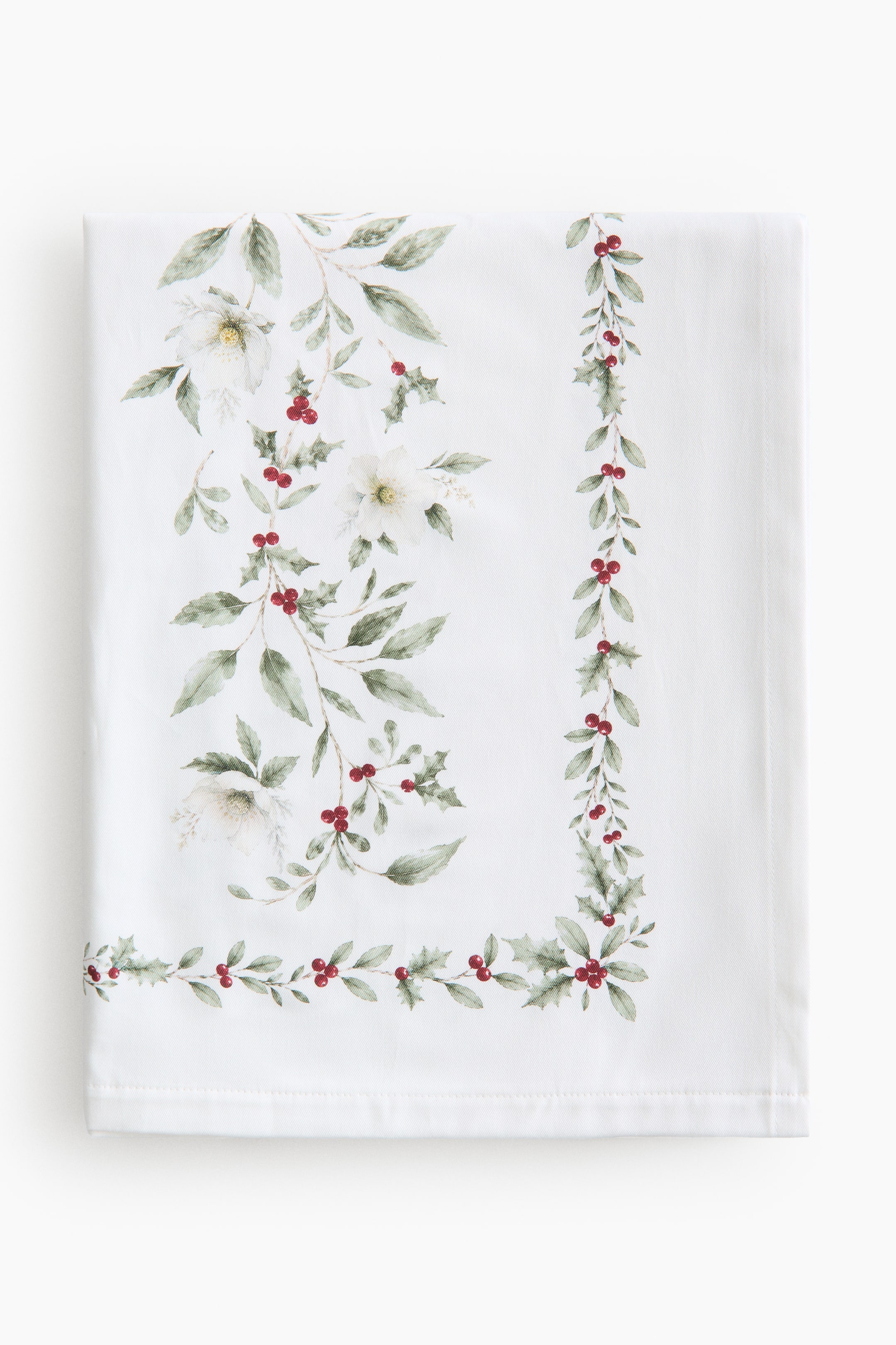 Agrandir l'image: Une nappe blanche pliée met en valeur, de face, son motif botanique d’angle, composé de feuilles vertes, de fleurs blanches et de grappes de baies rouges. Une délicate guirlande assortie de baies et de feuilles court le long de l’ourlet surpiqué.