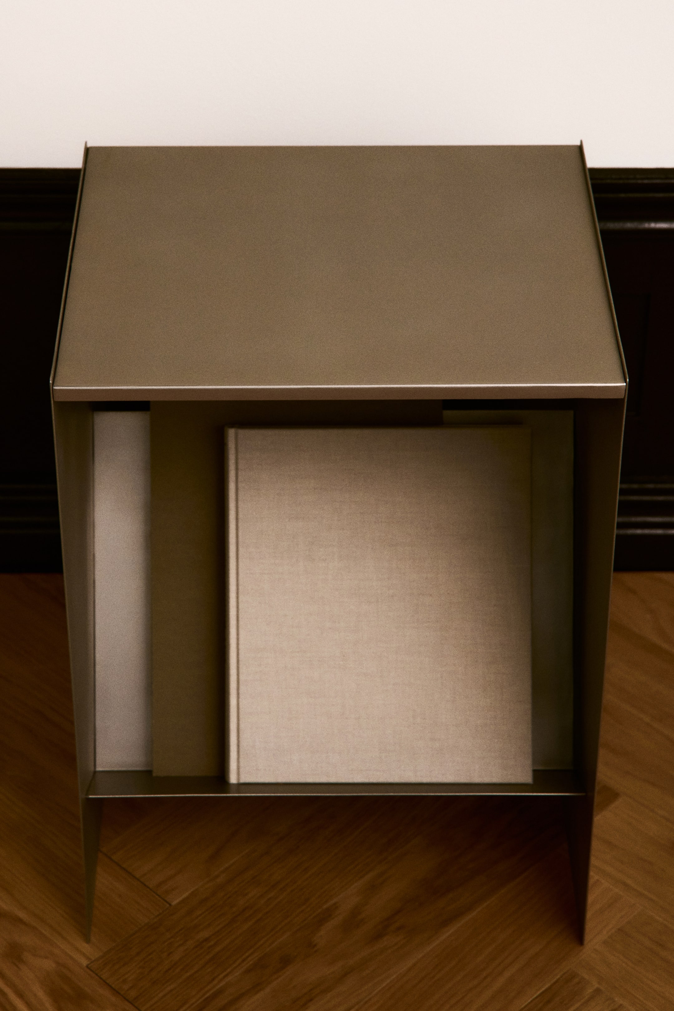 Ver imagem maior: Uma mesa lateral em metal de cor bronze, com um design geométrico, vista de frente, apresentando um tampo retangular plano e uma prateleira inferior integrada que segura um livro quadrado revestido em tecido.