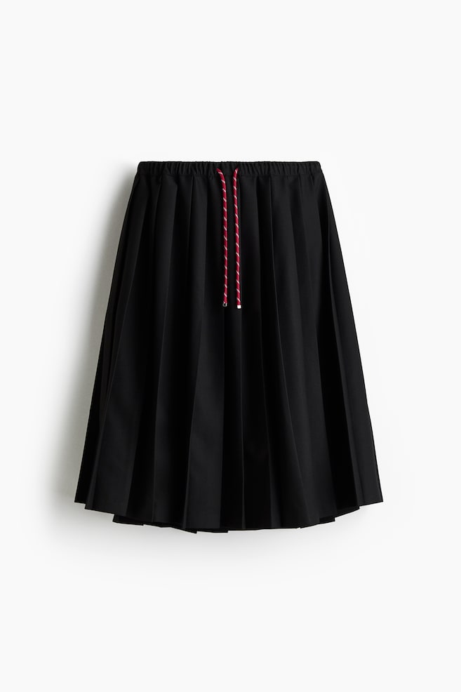 A black pleated skirt is shown from the FRONT, featuring an elasticated waistband. A red and white striped drawstring with silver-toned aglets is visible at the center front of the waistband. - 1