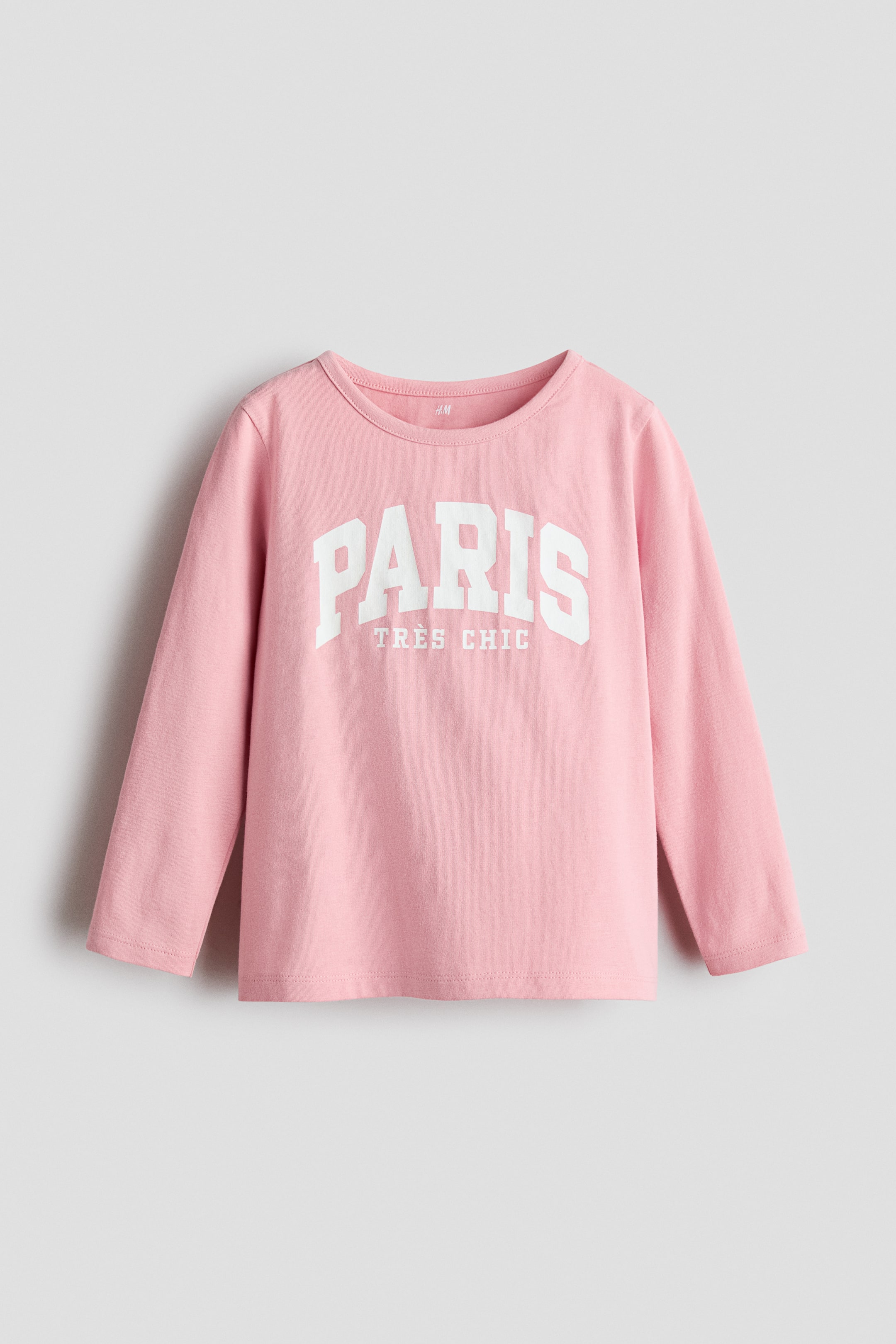 View larger image: A pink long-sleeve t-shirt is showcased from the front, featuring a round neck and long sleeves. The front of the t-shirt displays "PARIS" in large, arched white letters, with "TRÈS CHIC" in smaller white letters appearing just below.