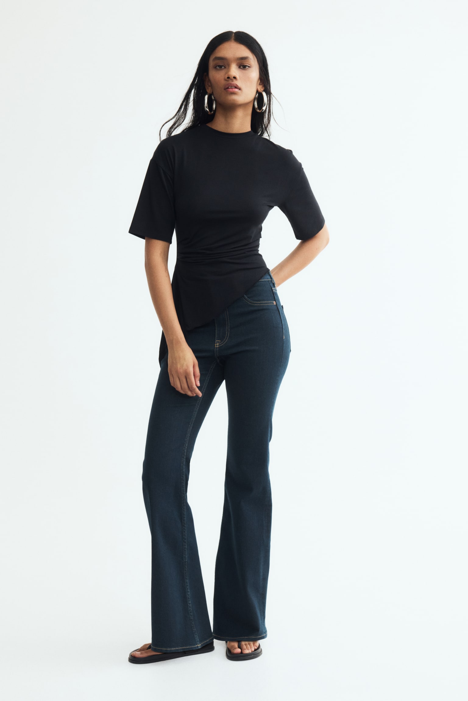 Dark blue denim jeans are shown front-facing and laid flat, featuring contrast orange stitching throughout. They feature a waistband with belt loops, a button closure with a zipper fly, front slash pockets, and a small coin pocket. The legs have a flared cut, widening from the hip to the hem. - 1