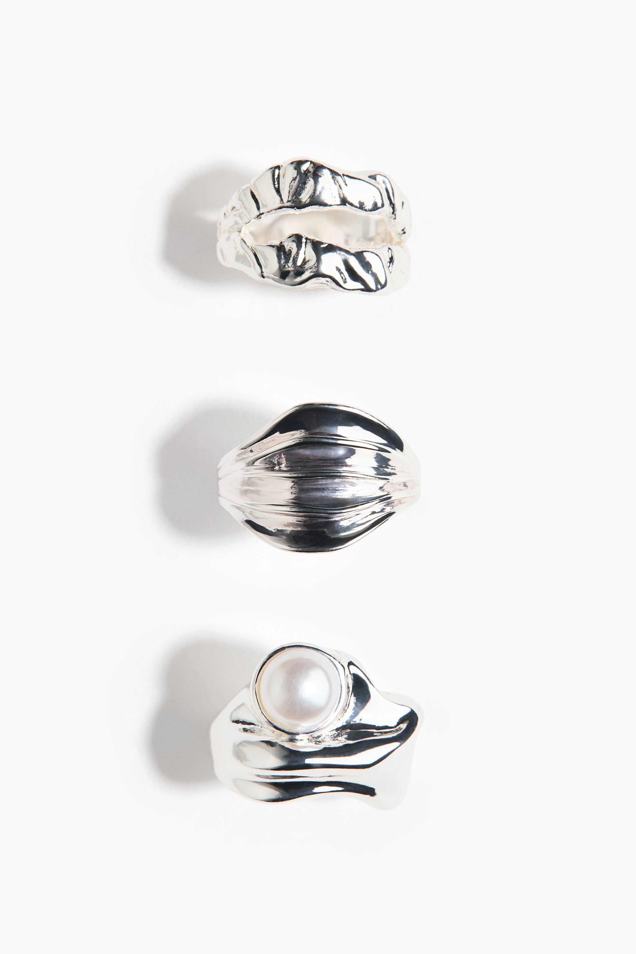 View larger image: Three silver-toned rings are lying flat, each displaying a unique design. The top ring is featuring a wide, organic, and textured surface, the middle ring is displaying a smooth, horizontally ribbed design, and the bottom ring is presenting a round, off-white pearl set centrally on a wide, irregular band.