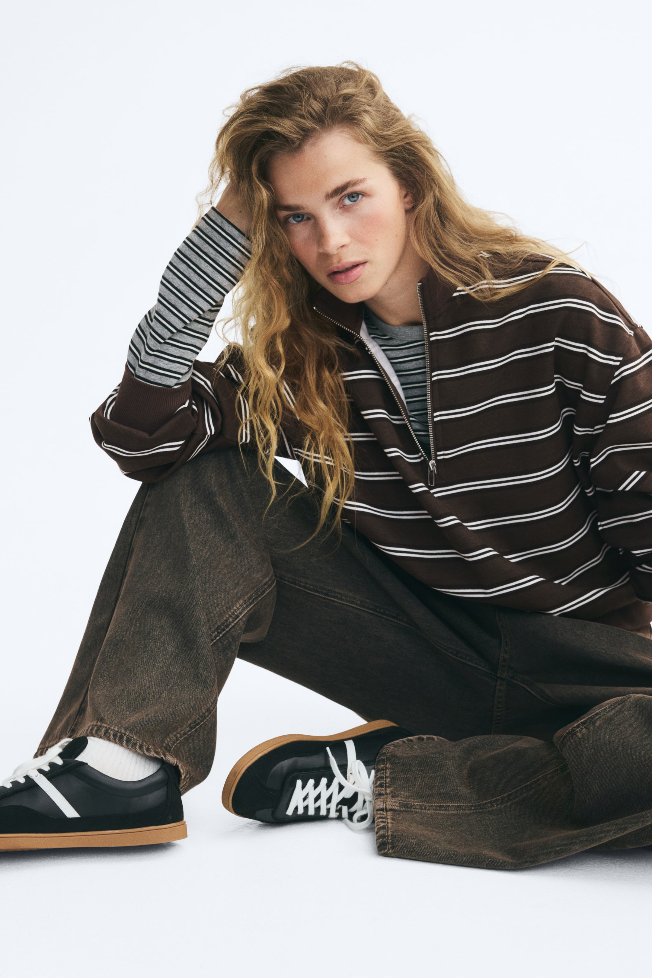View larger image: A brown and white horizontally striped half-zip sweater is being worn by the model, who is seated. The sweater features a stand collar with an open zip, dropped shoulders, and ribbed cuffs, and is layered over a grey striped long-sleeve shirt. It is styled with brown wide-leg jeans and black sneakers.