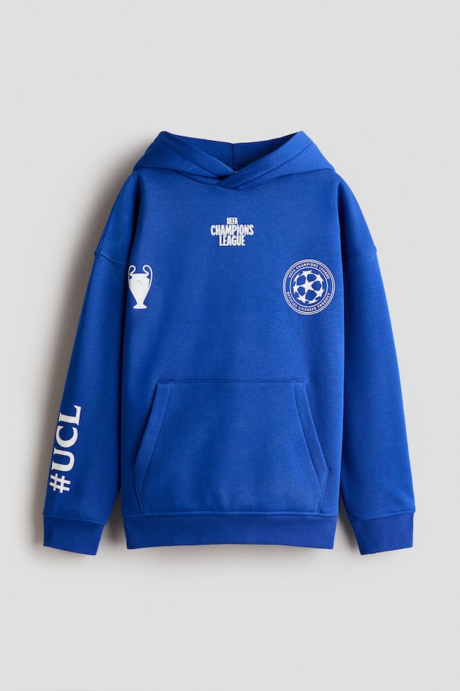 A blue sweat fabric hoodie is shown from the front, featuring a wrap-front hood, dropped shoulders, a kangaroo pocket, and ribbed trims at the cuffs and hem. The hoodie is detailed with white printed graphics, including UEFA CHAMPIONS LEAGUE text, a trophy icon, a circular logo, and #UCL printed vertically on the sleeve. - 1