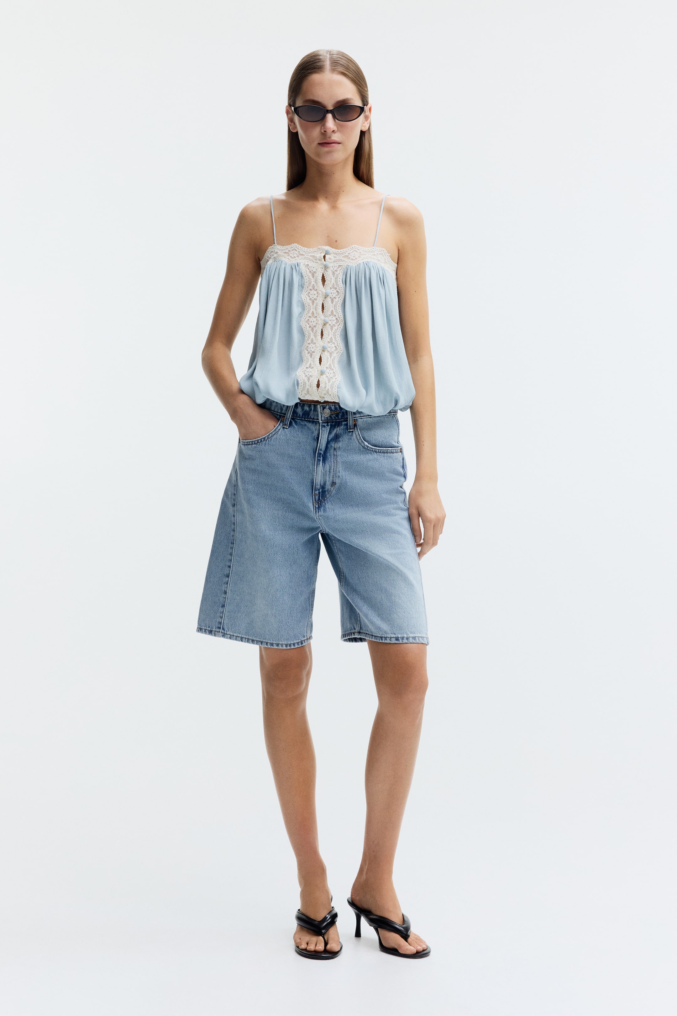 Grotere afbeelding bekijken: Knie-lange, 5-pocket shorts in licht gewassen, stijf katoenen denim worden gedragen door het model. De shorts zijn ontworpen met een hoge taille, een zichtbare ritssluiting en een knoopsluiting, en het model steekt één hand in de linker voorzak.