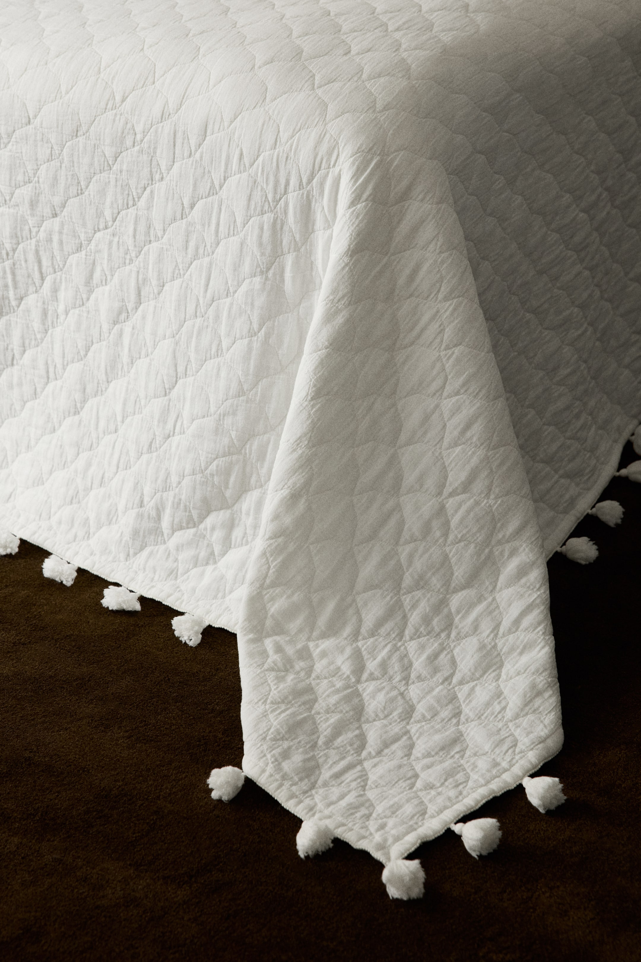 Agrandir l'image: Une couverture matelassée blanche présente un motif géométrique matelassé avec une texture froissée. Elle est bordée de petits pompons blancs le long de ses bords, et un coin est drapé vers le bas.