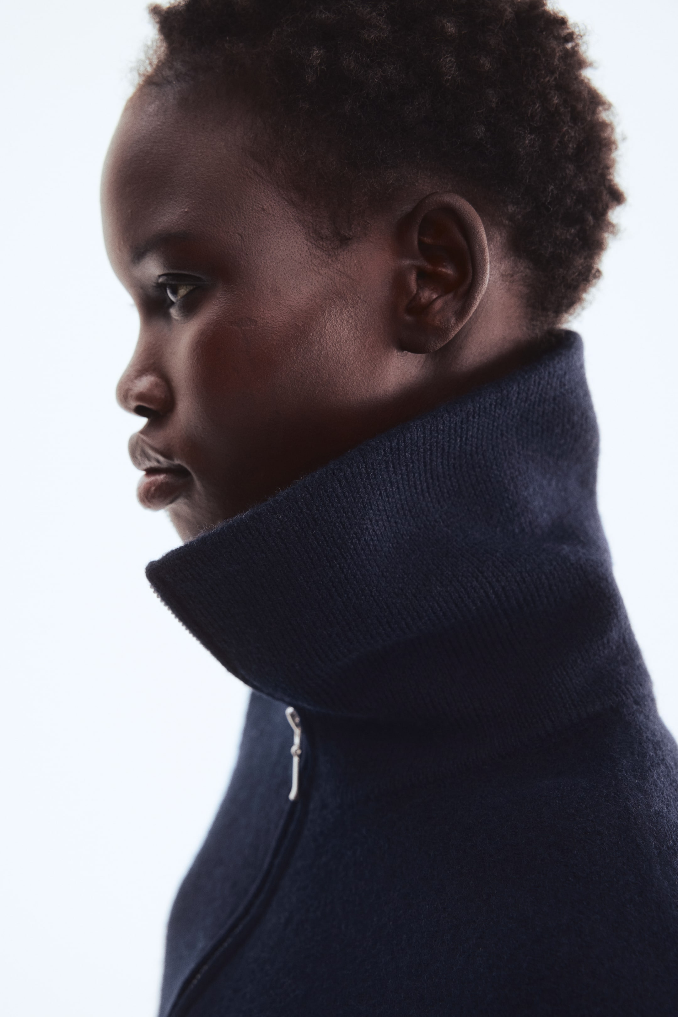 Visualizza immagine più grande: Un maglione blu navy scuro presenta un alto colletto a coste e una zip parziale color argento, con il tessuto testurizzato visibile da un profilo laterale.