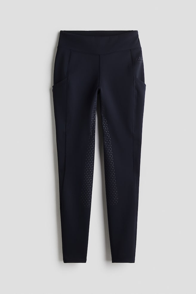 Ein Paar dunkelblaue Reitleggings wird von vorne präsentiert, mit einem breiten Bund und tiefen Seitentaschen an beiden Oberschenkeln. Die Innenseiten der Oberschenkel und Knie zeigen ein strukturiertes Griffmuster, und ein dezentes tonales Logo ist auf dem oberen rechten Oberschenkel sichtbar. - 1