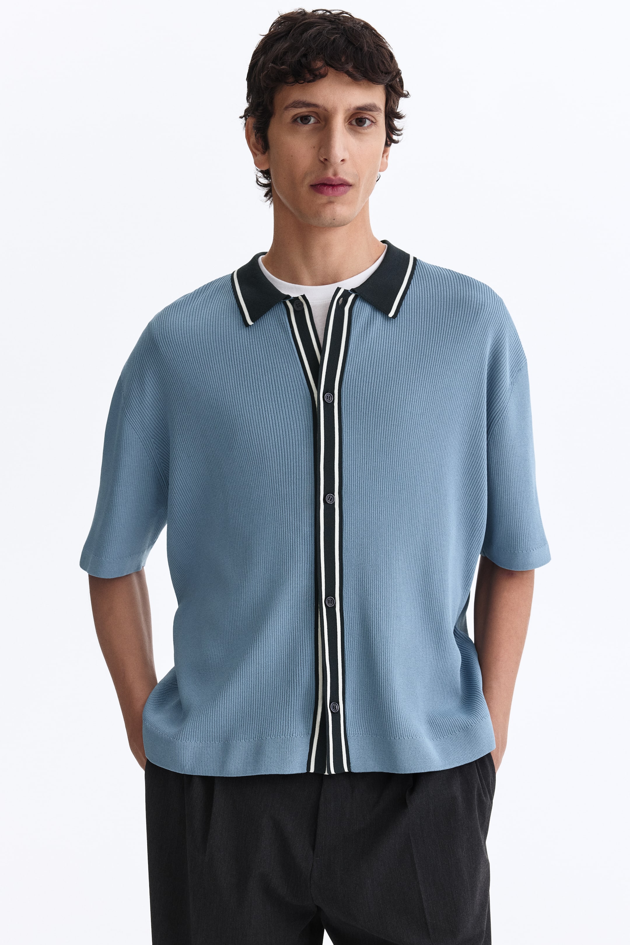 Ampliar la imagen: Una camisa azul de manga corta con cuello, mostrada de frente, presenta una textura acanalada, un cuello spread negro con una franja blanca a lo largo del borde y una tapeta con botones a todo lo largo con ribete negro y dos franjas blancas paralelas; se lleva abierta sobre una camiseta blanca.