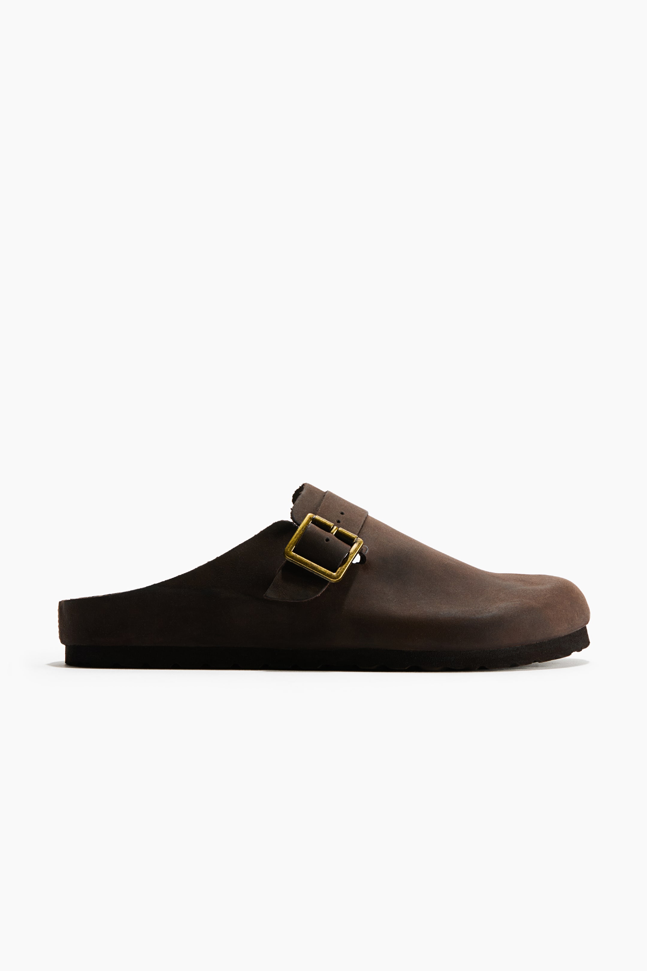 View larger image: A dark brown leather mule is shown from a side profile view, featuring a closed, rounded toe and an open back. The mule has an adjustable strap across the instep with a rectangular brass-toned metal buckle, resting on a flat, dark sole with a patterned tread.