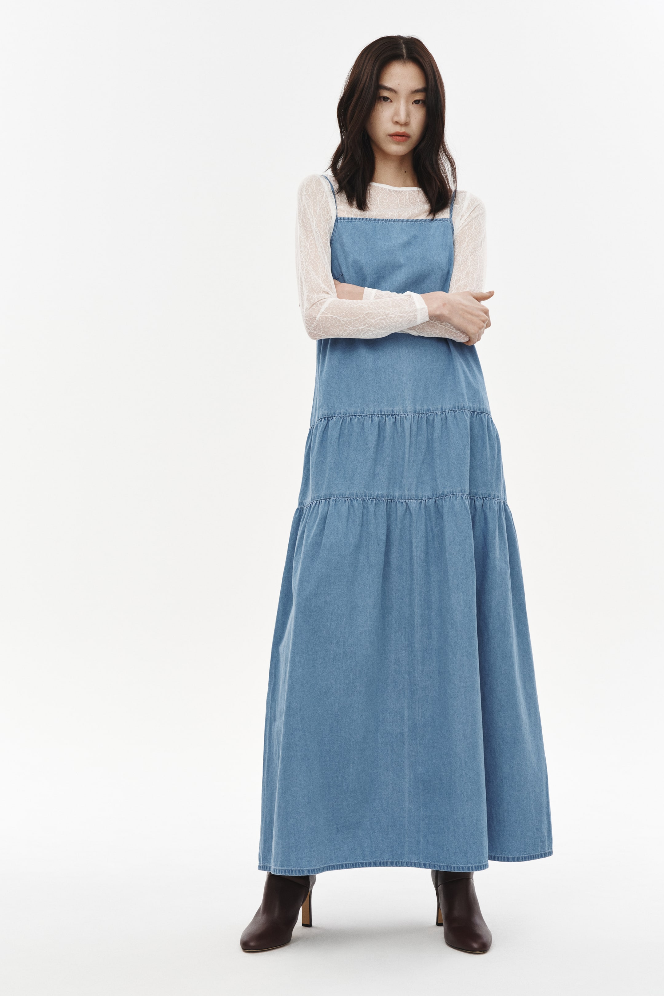 拡大画像を表示: ライトブルーのデニムマキシドレスは、前面から見たデザインで、ギャザー入りのティアードスカートと細いショルダーストラップが特徴です。白の長袖レーストップ、ラウンドネックラインと重ね着されています。