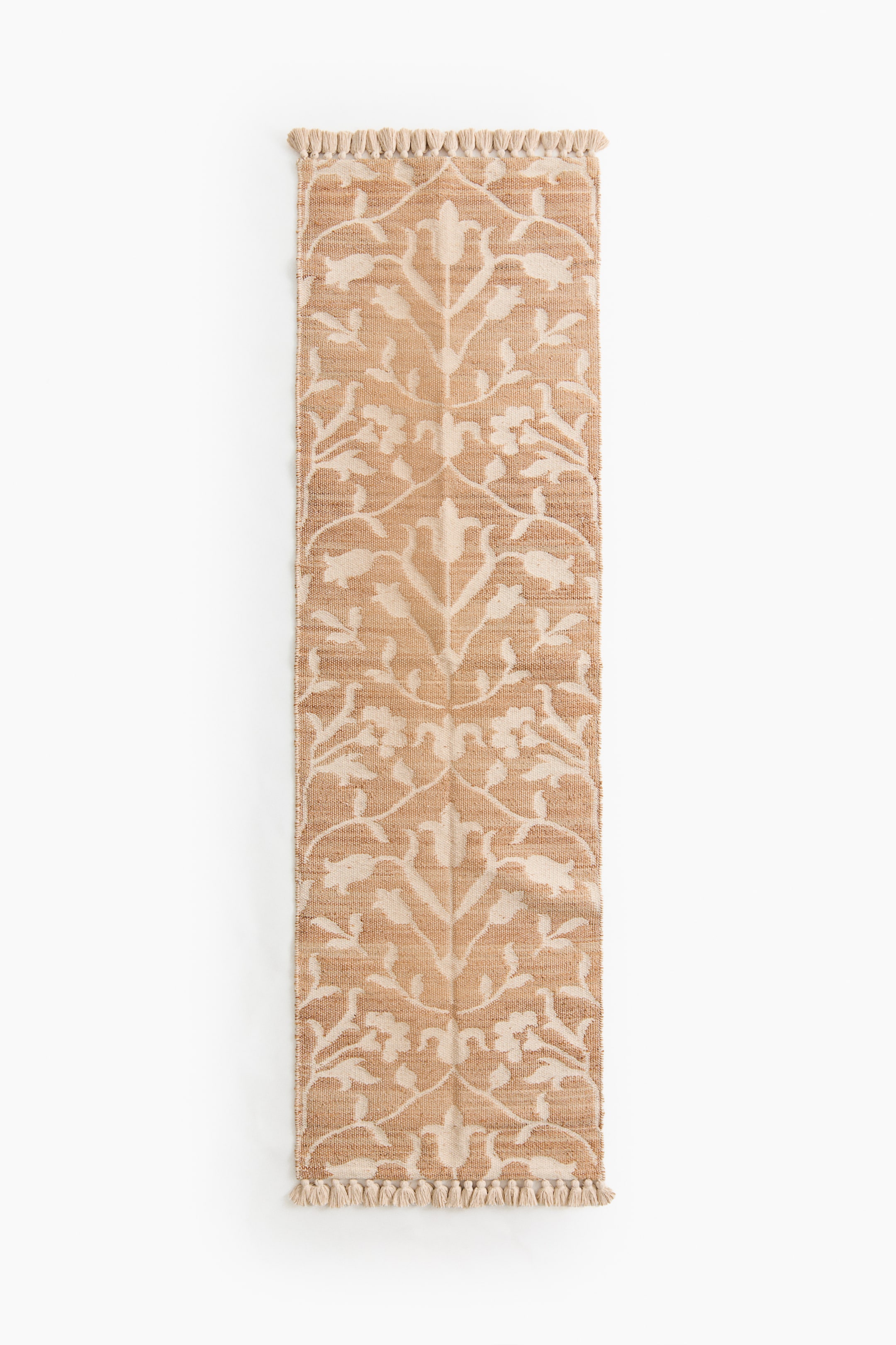 View larger image: A rectangular runner rug displays a symmetrical floral-and-vine pattern in light beige against a tan woven background. Short fringes adorn both ends of the rug.