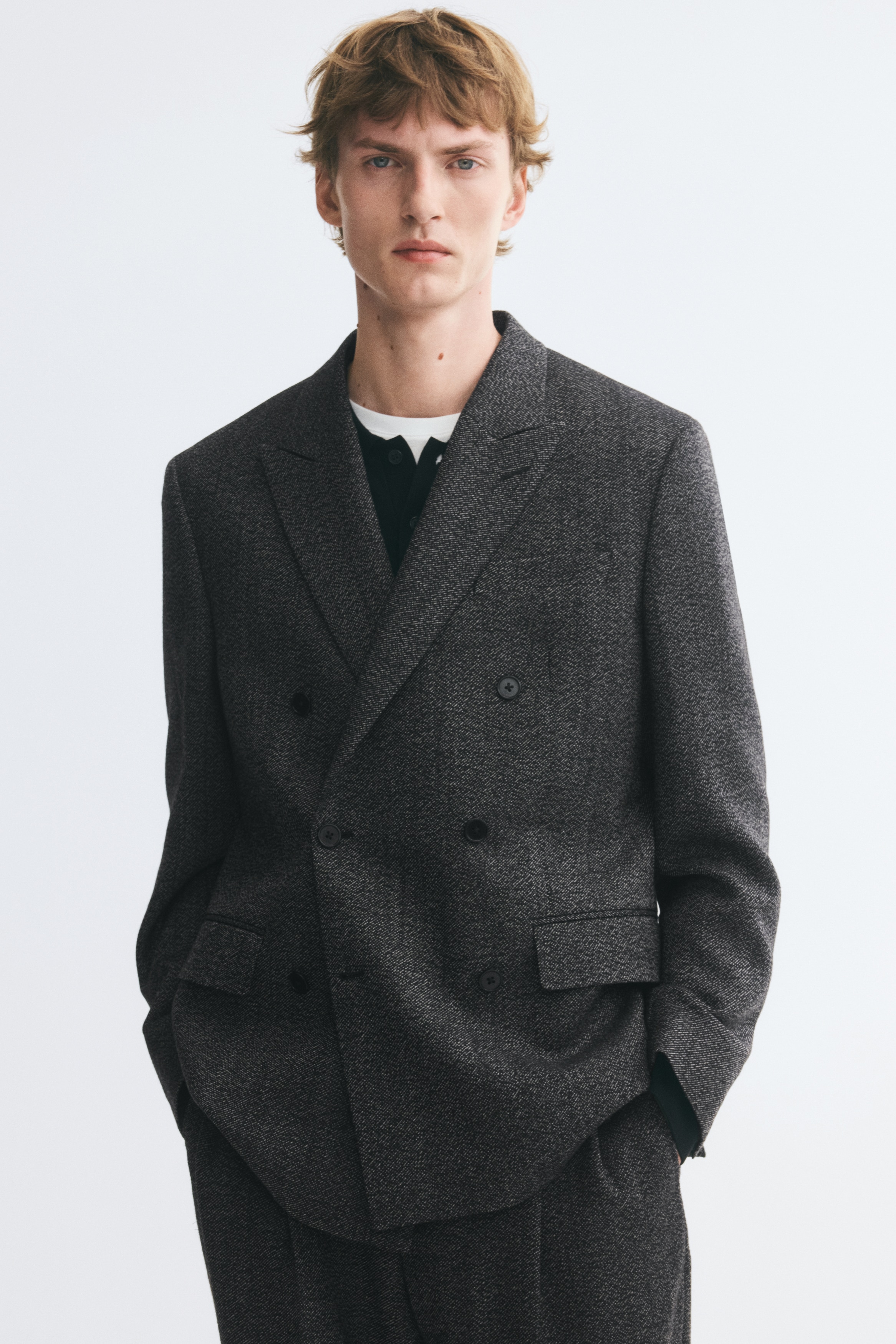 A dark grey, textured double-breasted blazer is shown from the front, featuring wide peak lapels and a six-button front fastening. The structured jacket features two lower flap pockets and a subtle speckled weave throughout the fabric.