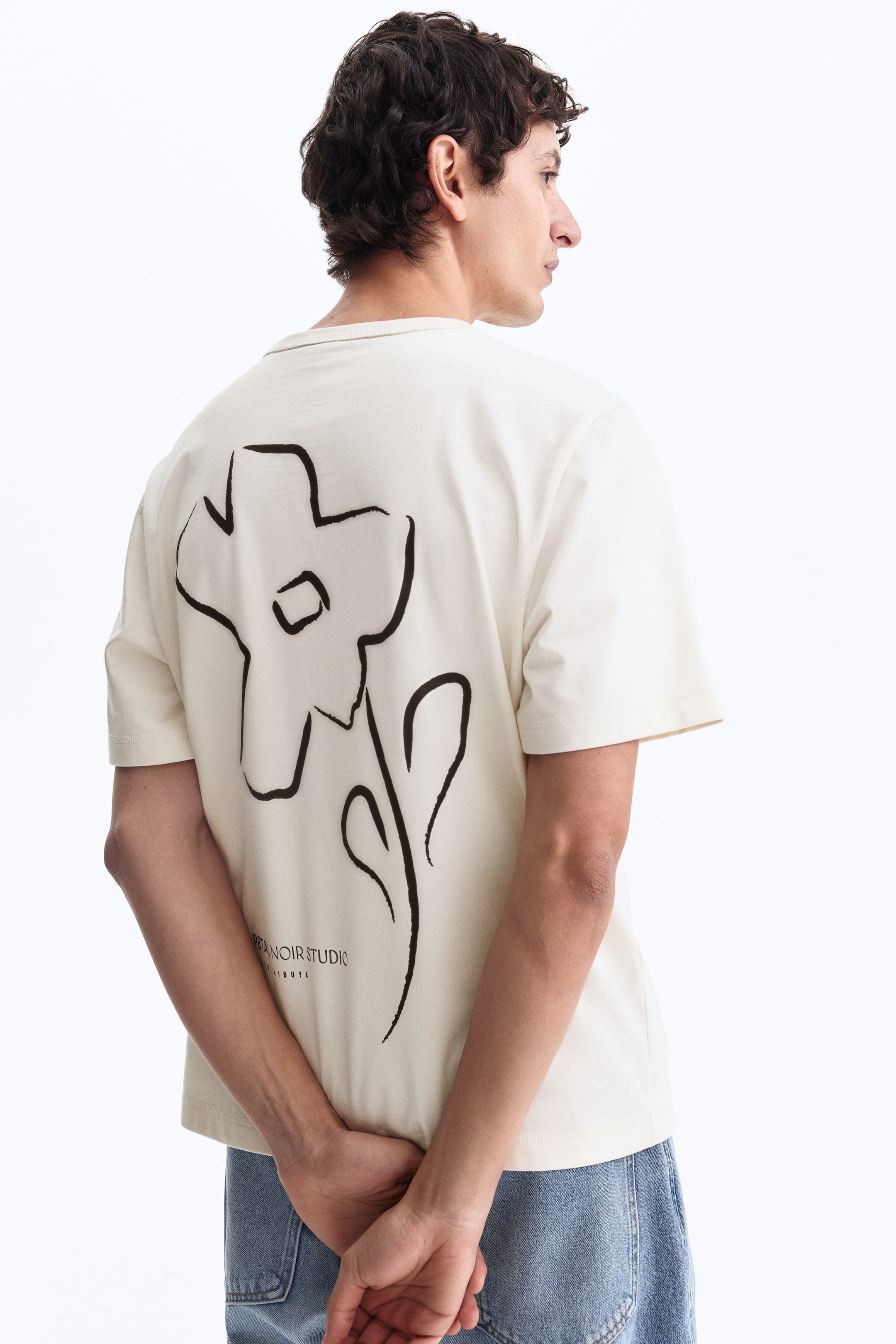 View larger image: A cream T-shirt shown from the back, featuring a large black outline of a flower. Below the flower, the text "BETA NOIR STUDIO LONDON" is printed.