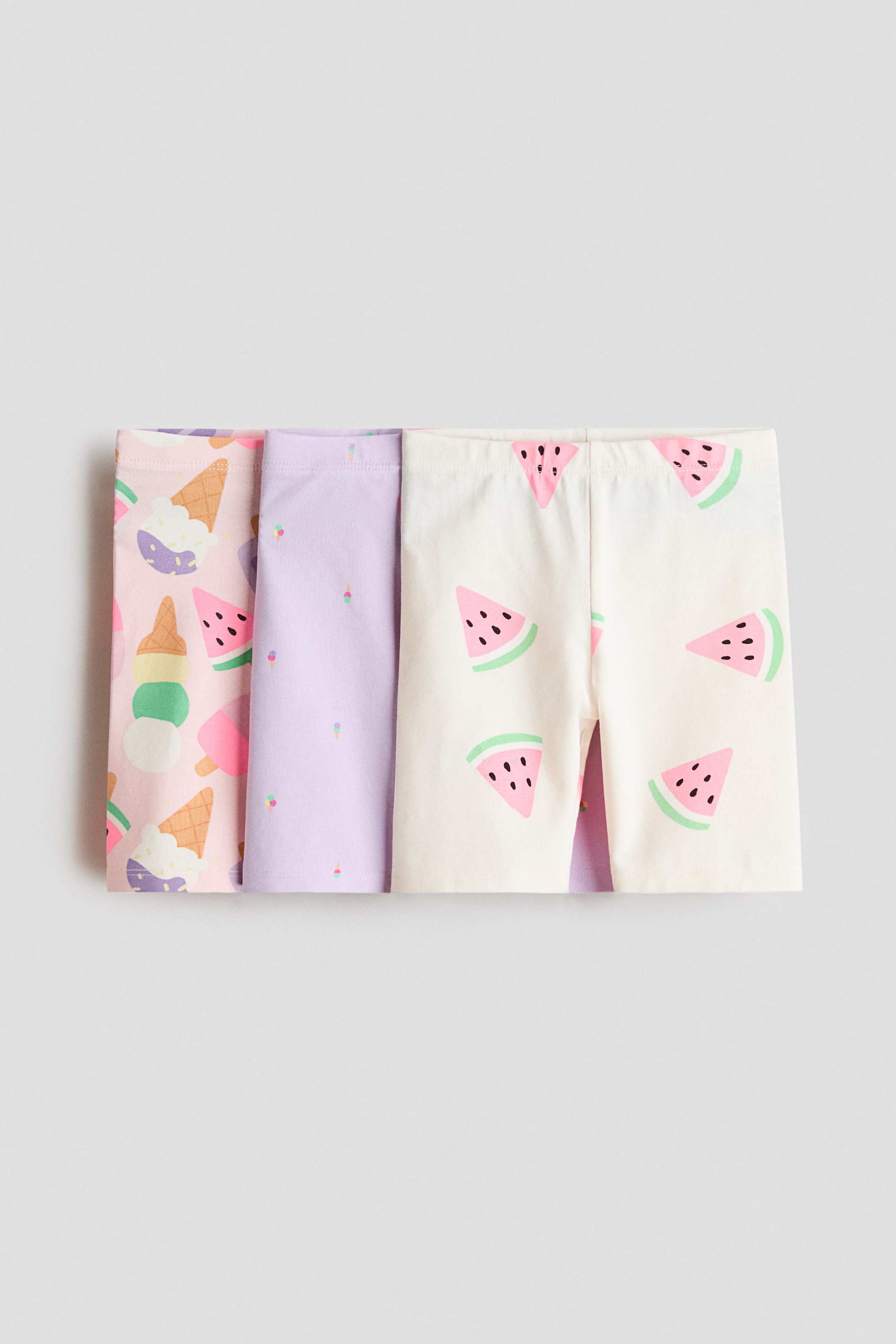 Agrandir l'image: Trois paires de shorts de cyclisme sont pliées et empilées, présentant leur vue de face. Chaque paire est confectionnée en jersey de coton doux, dotée d'une taille élastiquée, et arbore un motif unique : une paire rose avec des cornets de glace et des tranches de pastèque, une paire lavande avec de petits cornets de glace, et une paire blanc cassé avec des tranches de pastèque.