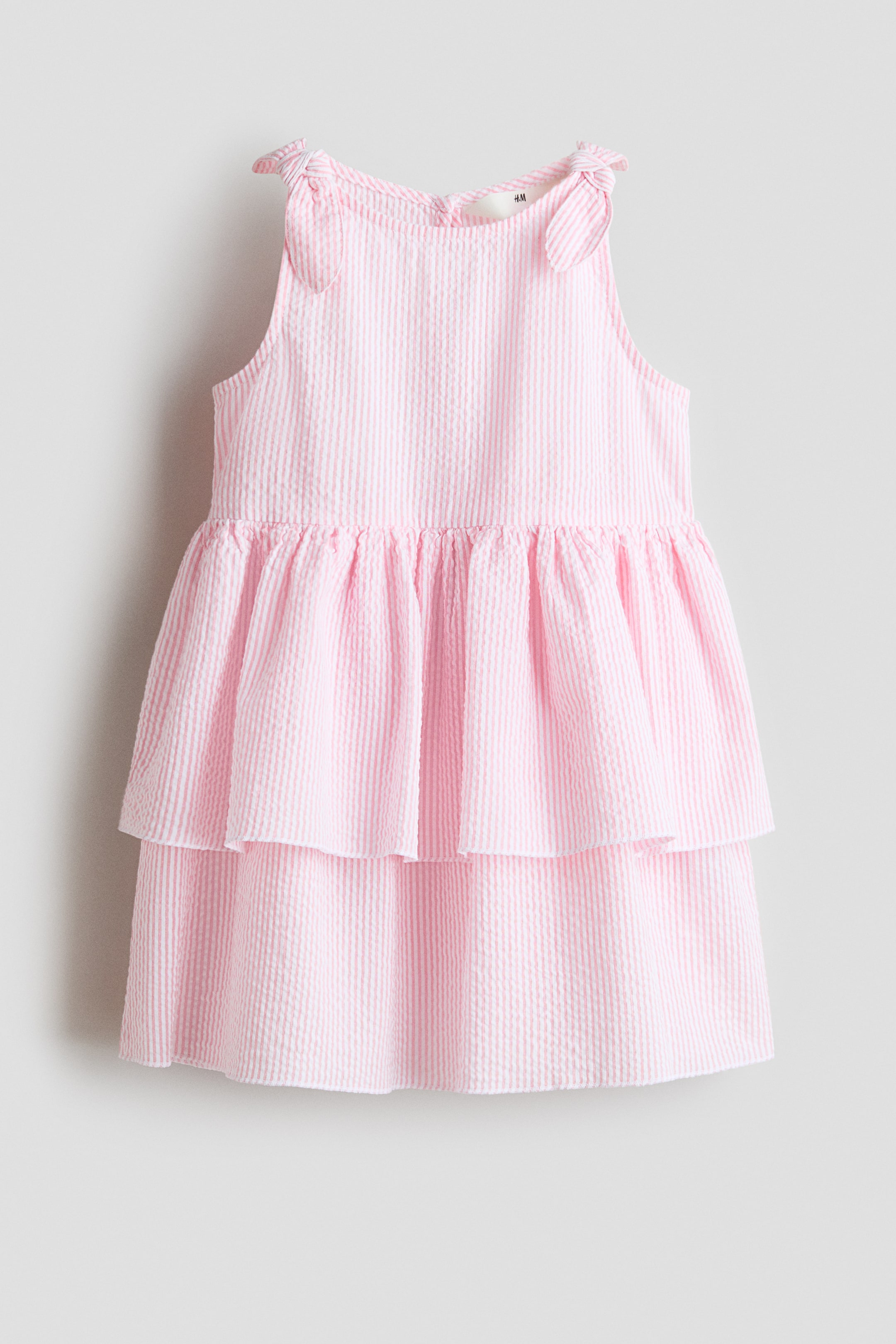 View larger image: A pink-and-white striped seersucker dress shows its front, featuring a rounded neckline and shoulder straps tied in bows. It has a gathered waistline and a two-tiered skirt, with both tiers gathered.