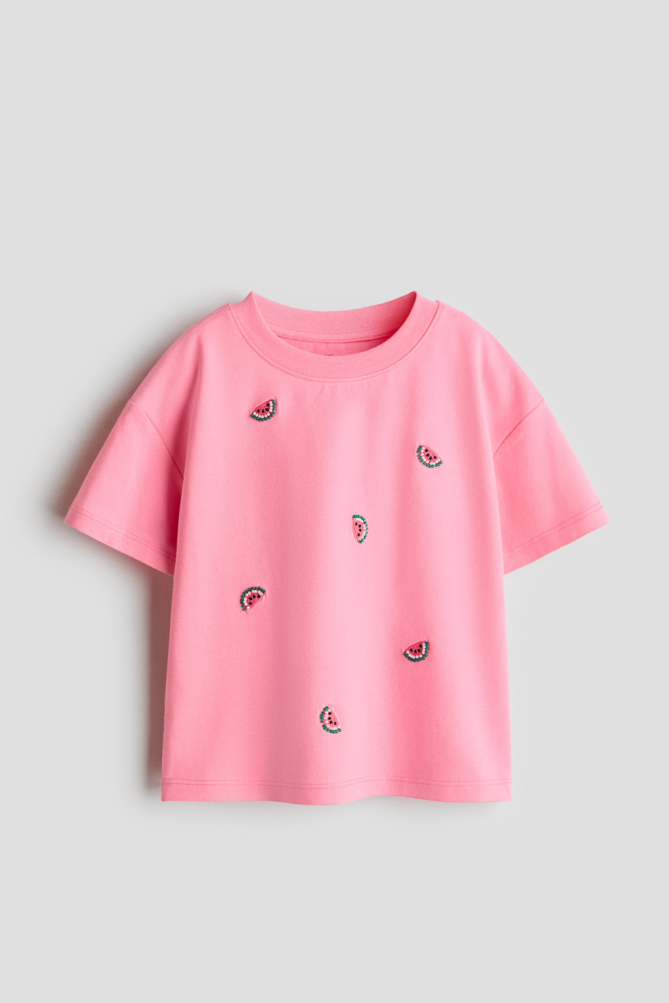 View larger image: A relaxed-fit, pink T-shirt is showcasing its front, featuring a ribbed trim around the neckline and low dropped shoulders. It is adorned with scattered watermelon slice designs.