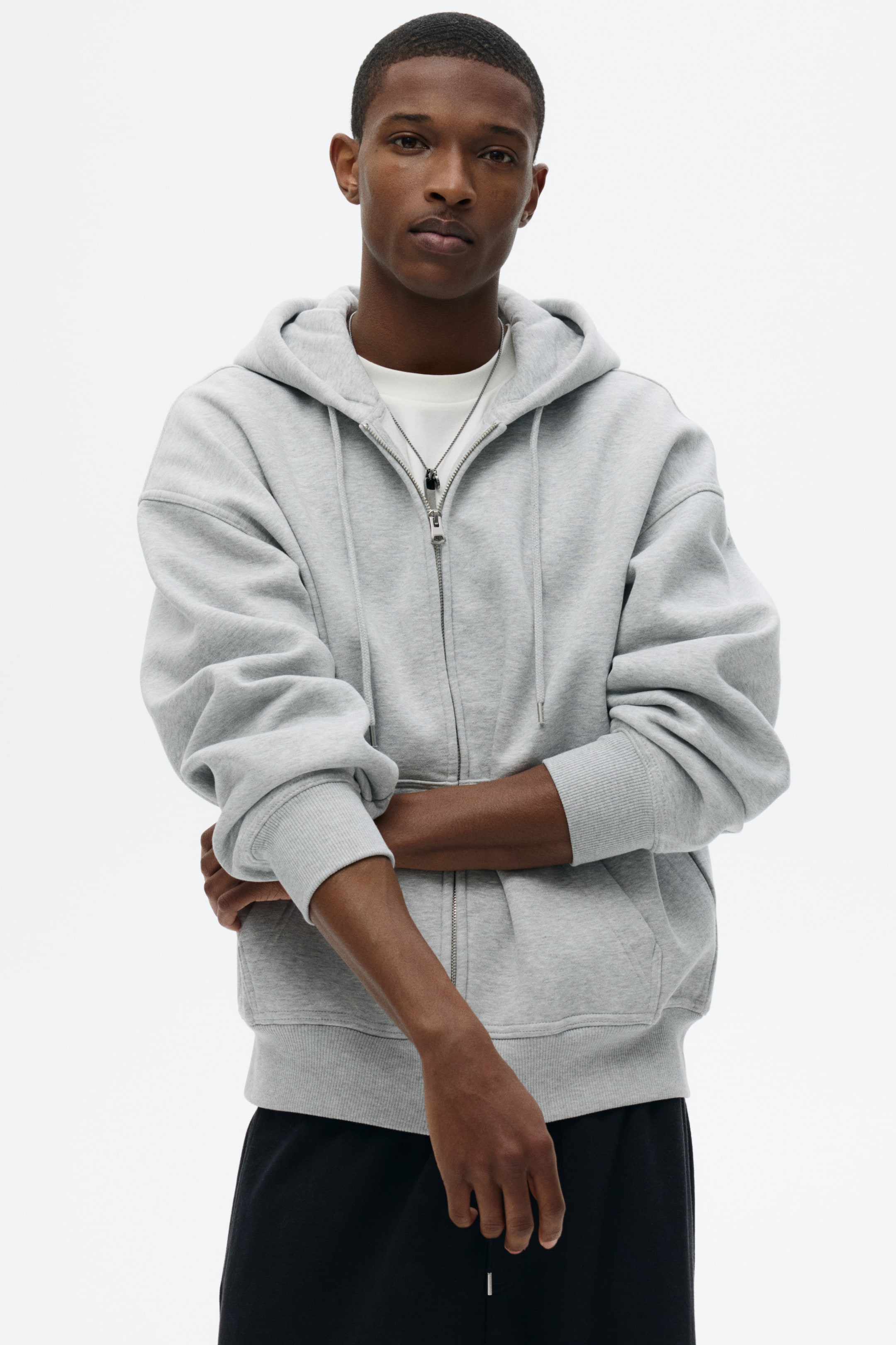 View larger image: A grey full-zip hoodie is presented from a front-facing angle, featuring a silver-toned zipper that is partially open, revealing a white crew-neck T-shirt and a silver necklace. The hoodie showcases a relaxed fit with dropped shoulders, ribbed cuffs, a ribbed hem, and visible drawstrings on the hood.