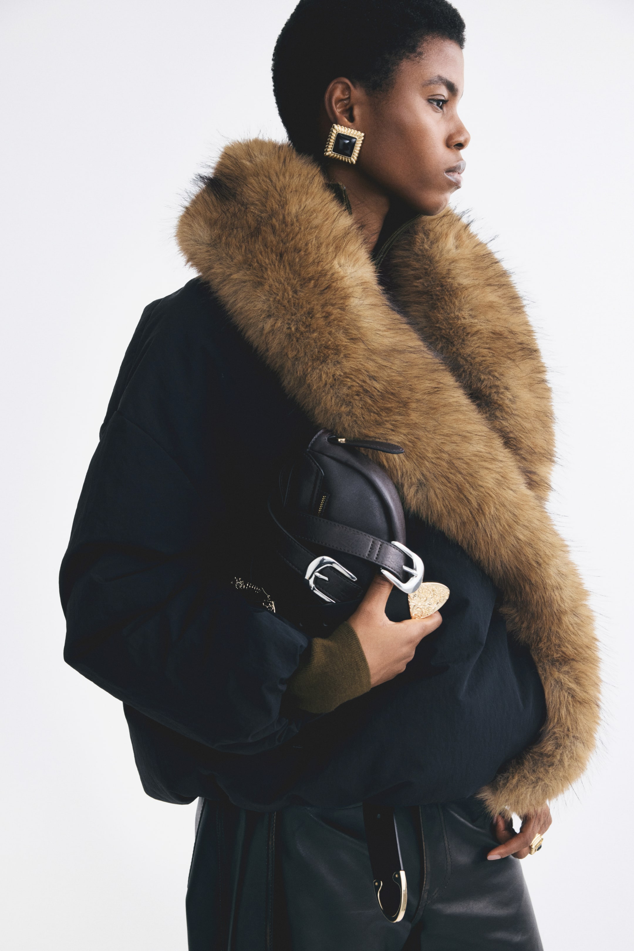 View larger image: An oversized, padded black nylon jacket featuring a wide brown faux-fur shawl collar is being worn by the model. The model is holding a black leather handbag with silver buckles against the front of the jacket, which is styled with dark leather trousers.