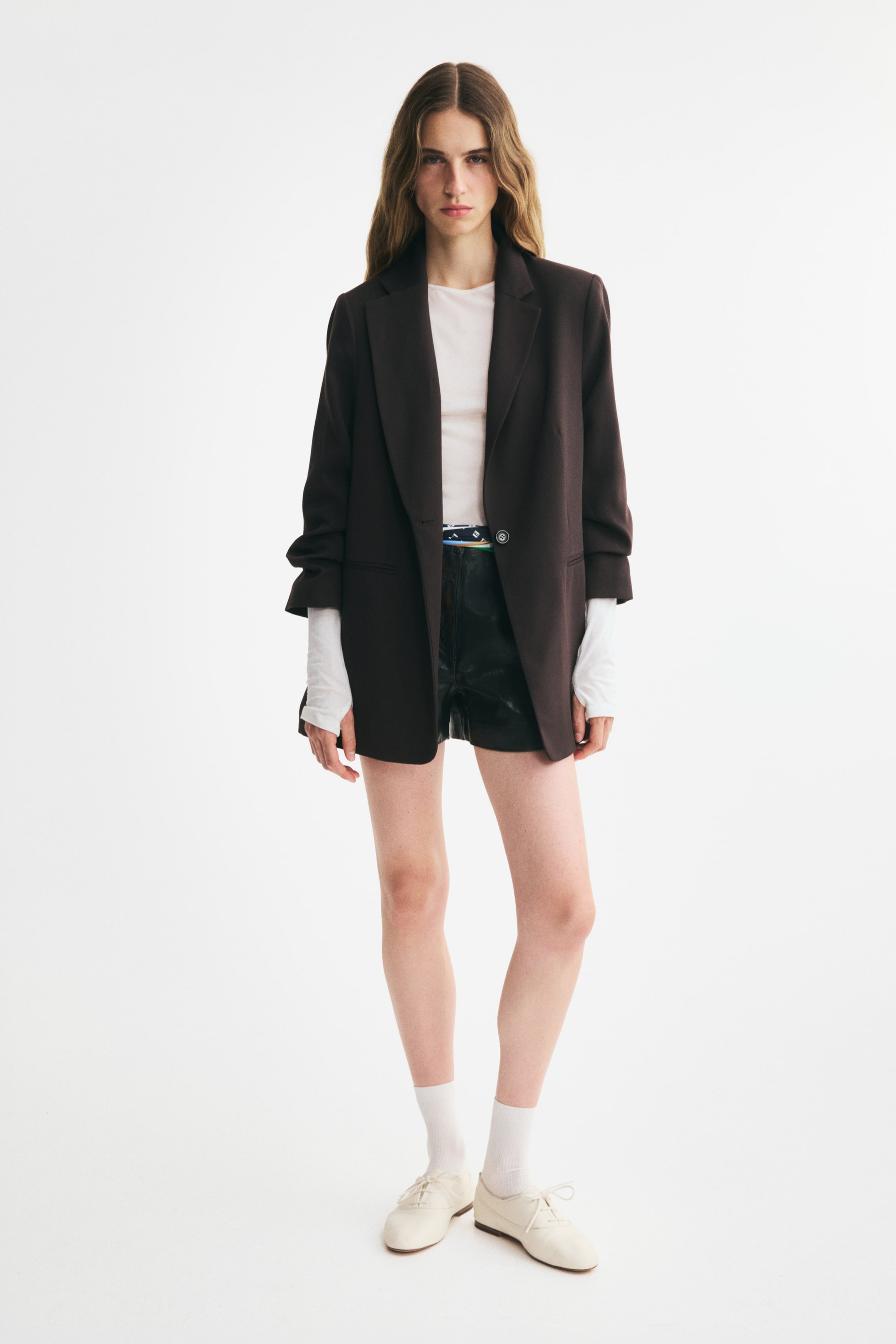 View larger image: A long, single-breasted blazer in dark brown woven fabric is shown from the front, featuring notch lapels, a single-button closure, and gathered 3/4-length sleeves. The blazer is styled open over a white long-sleeve top and black shorts, highlighting its relaxed fit and structured shoulder pads.