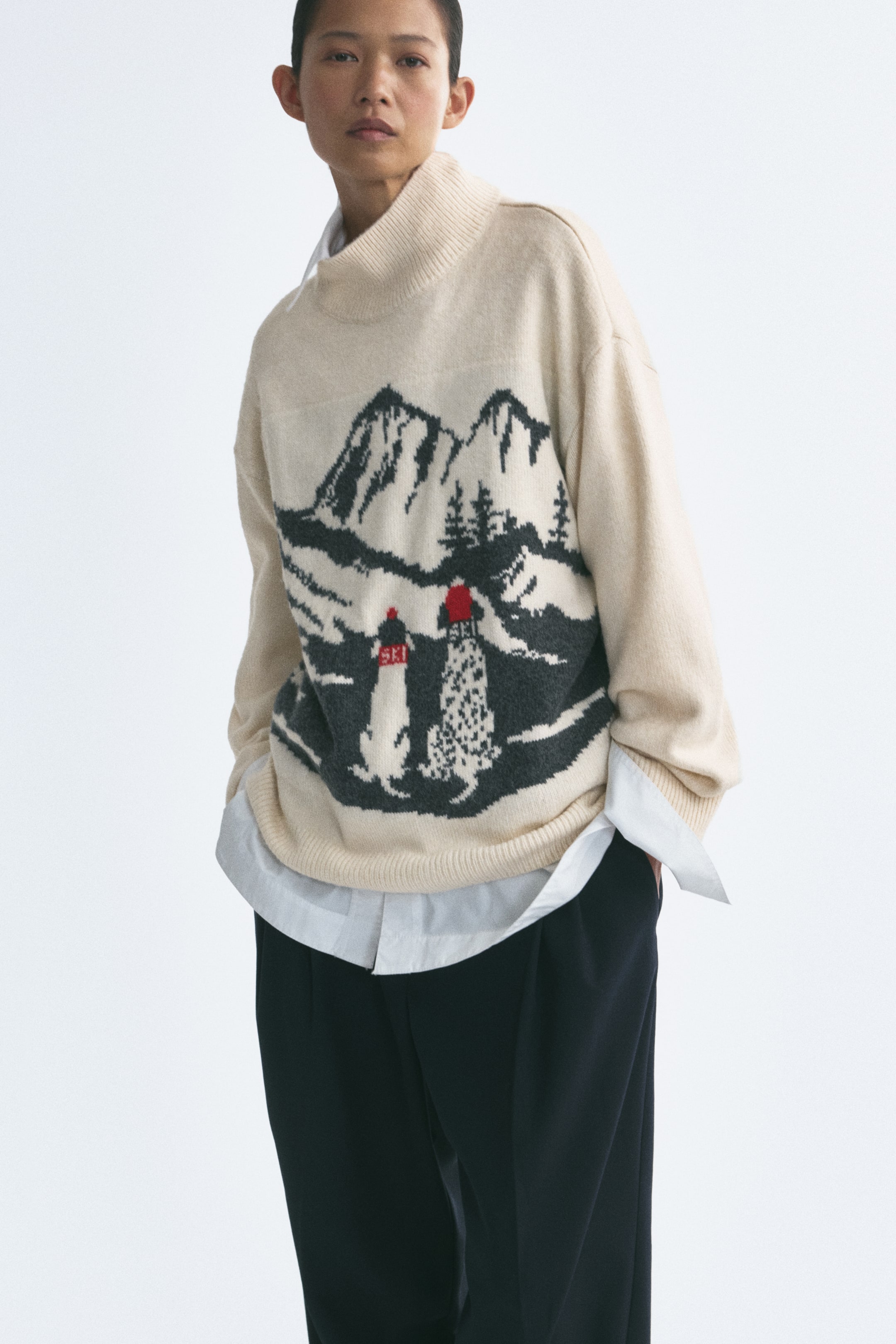 Ampliar la imagen: La modelo lleva un suéter de punto jacquard color crema con cuello alto acanalado, hombros caídos y un gráfico de paisaje montañoso en negro y rojo. El suéter se lleva sobre una camisa blanca con cuello y se combina con pantalones oscuros.