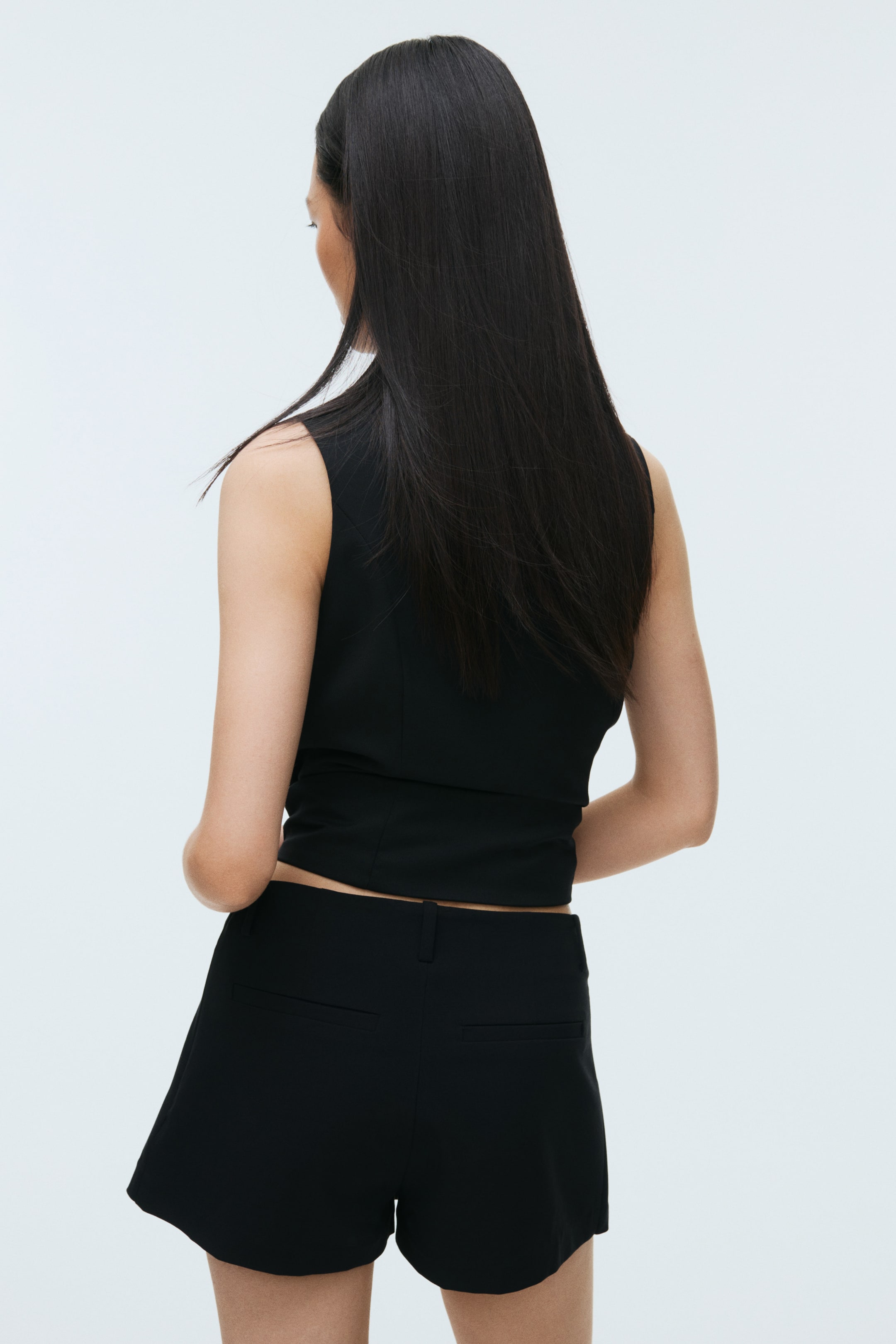 Ampliar la imagen: Un top corto negro sin mangas presenta una espalda ajustada con costuras verticales, terminando justo por encima de la cintura. Los shorts negros a medida muestran bolsillos de ojal traseros y un dobladillo ligeramente curvado, con ambas prendas vistas desde atrás.