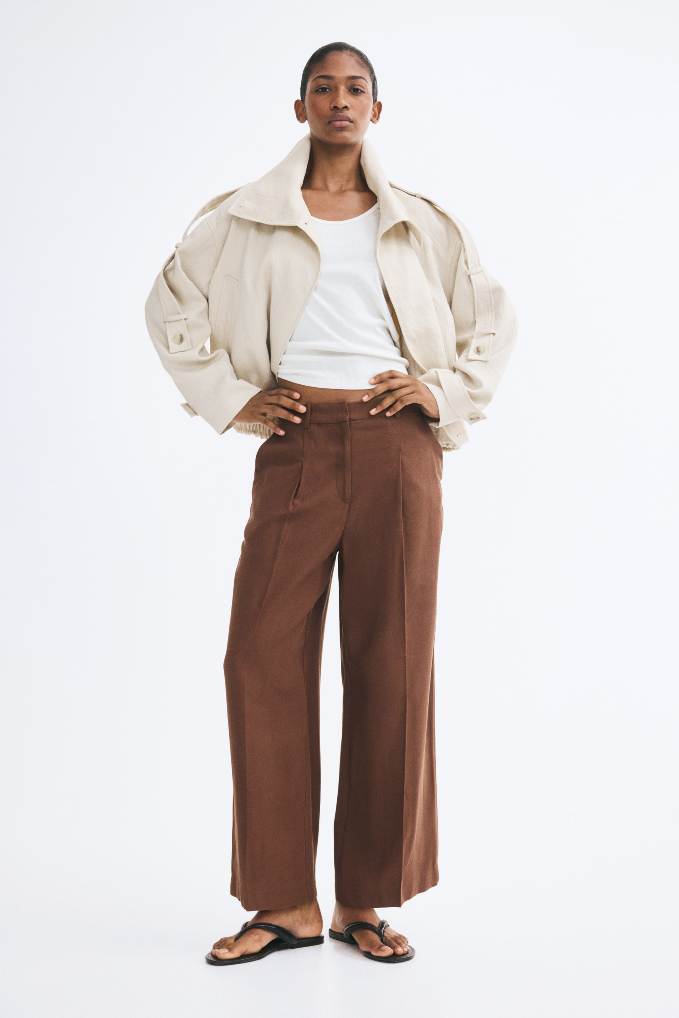 Ampliar la imagen: La modelo lleva unos pantalones café de tiro alto y pierna recta con dobleces marcados al frente, mientras posa de pie con las manos descansando cerca de la pretina. Los pantalones tobilleros están combinados con una blusa blanca, un saco color crema abierto y sandalias negras tipo thong.