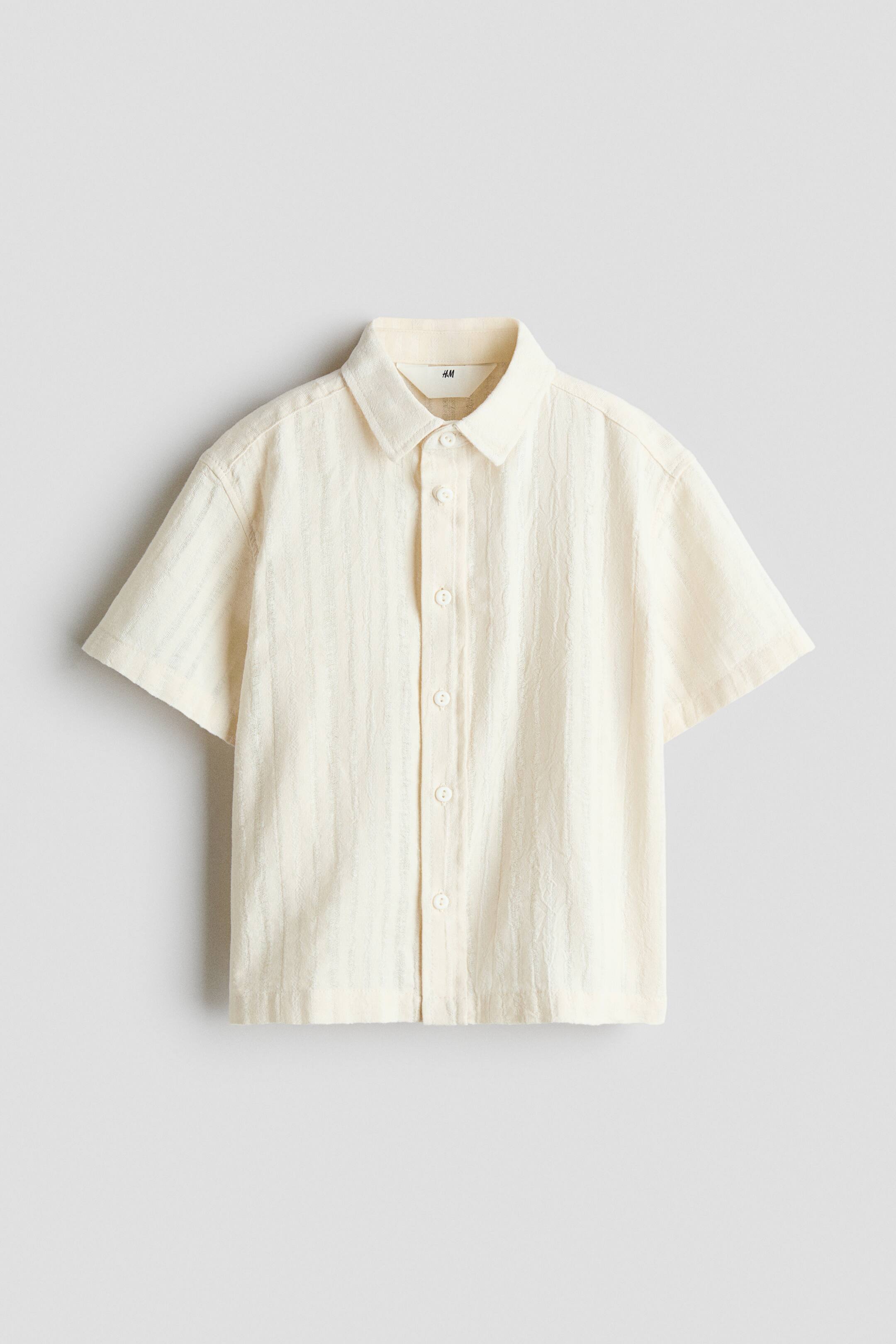 View larger image: A short-sleeved, off-white button-up shirt is shown from the front, displaying textured, vertically crinkled fabric. The shirt features a classic turn-down collar, a front button placket with matching white buttons, and a straight hem.