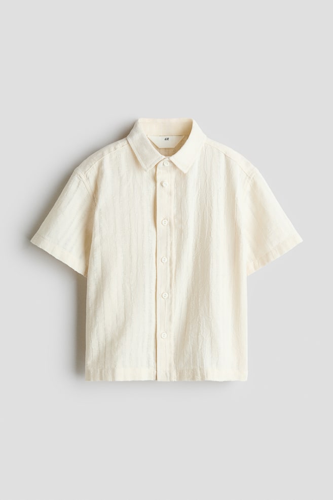 A short-sleeved, off-white button-up shirt is shown from the front, displaying textured, vertically crinkled fabric. The shirt features a classic turn-down collar, a front button placket with matching white buttons, and a straight hem. - 1