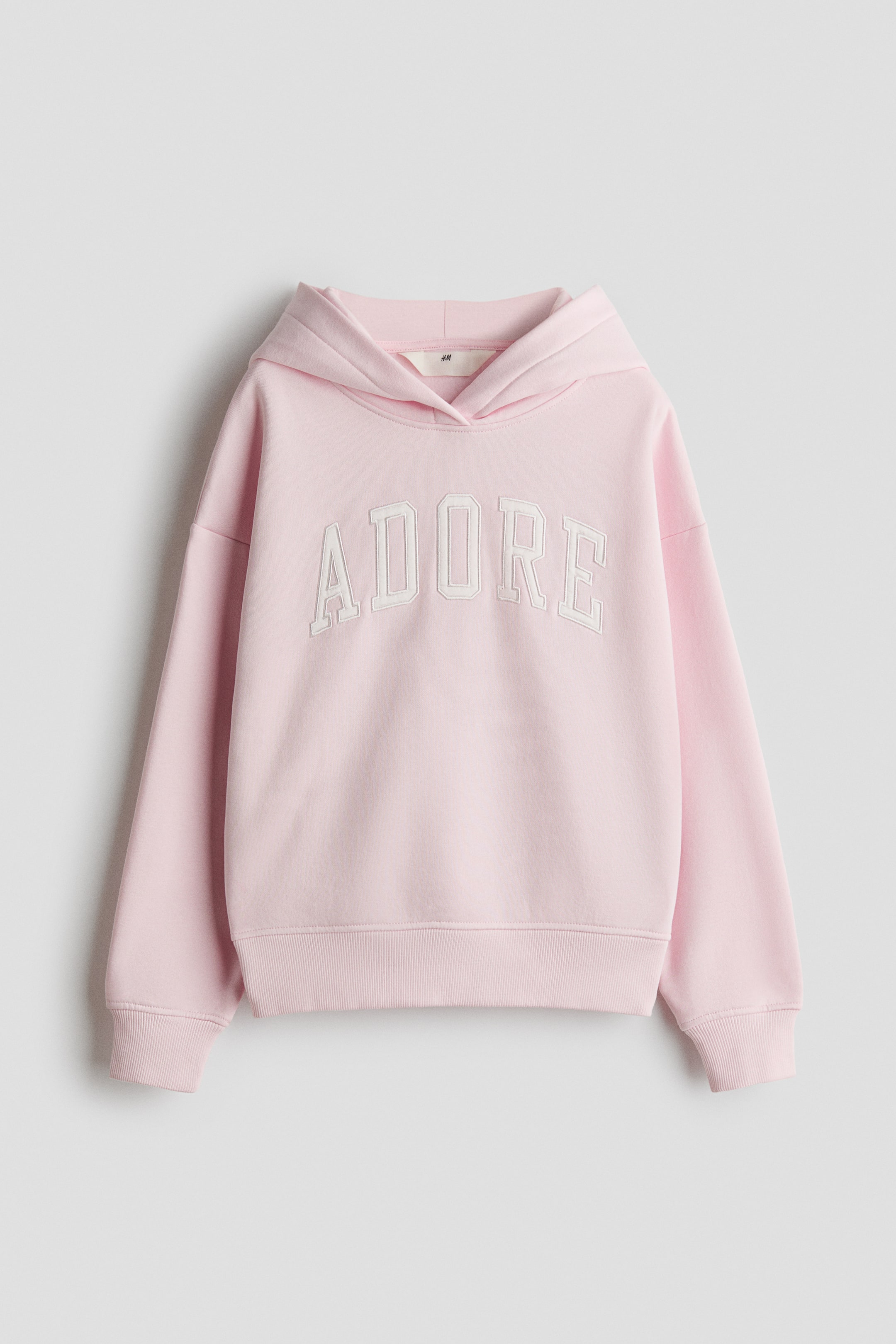 Vis større bilde: En rosa hettegenser viser ordet "ADORE" i hvite broderte bokstaver over brystet. Den avslappede hetten er synlig, og den har ribbestrikkede mansjetter og en ribbestrikket kant, presentert flatt og vendt fremover.
